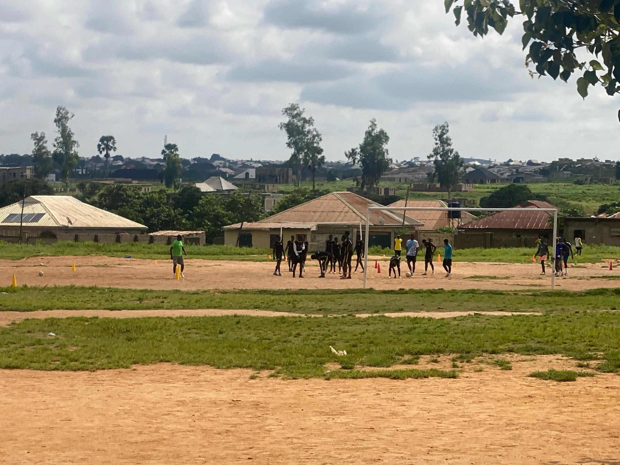 In Narayi, Nigeria, a group of young men are engaged in a football training session on a dusty, patchy field under an overcast sky. The field is marked with orange cones and a single football is visible near a group of players. In the background, several single-story buildings with corrugated iron roofs are scattered amongst trees and vegetation, suggesting a semi-urban or rural setting. A white goalpost stands on the right side of the frame, with several figures clustered around it. Another individual in a bright green shirt is visible further to the left, appearing to be training separately. The overall scene depicts a casual sporting activity taking place within a community.