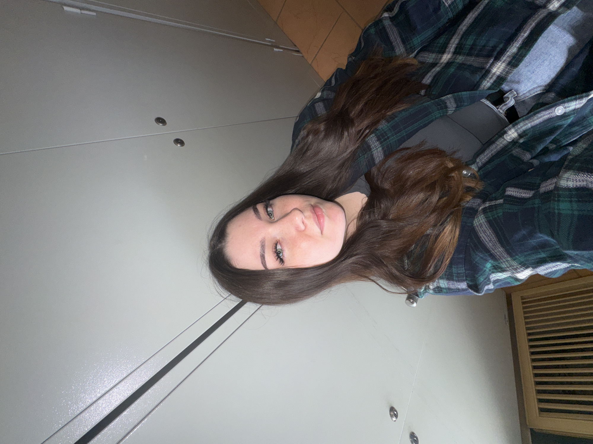 A young woman with long brown hair is taking a selfie in what appears to be a locker room or a similar facility. She is wearing a dark grey top, a green and blue plaid shirt unbuttoned over the top, and light-wash jeans. She has on light makeup and is looking directly at the camera with a slight smile. The background consists of a light-colored wall with horizontal accents and circular fixtures, suggestive of a public or institutional setting. The floor appears to be tiled with orange and brown square tiles. Based on the user's request to place the scene in Perm, Russia, this could be a locker room at a sports facility, a swimming pool, or a public changing room in that city.