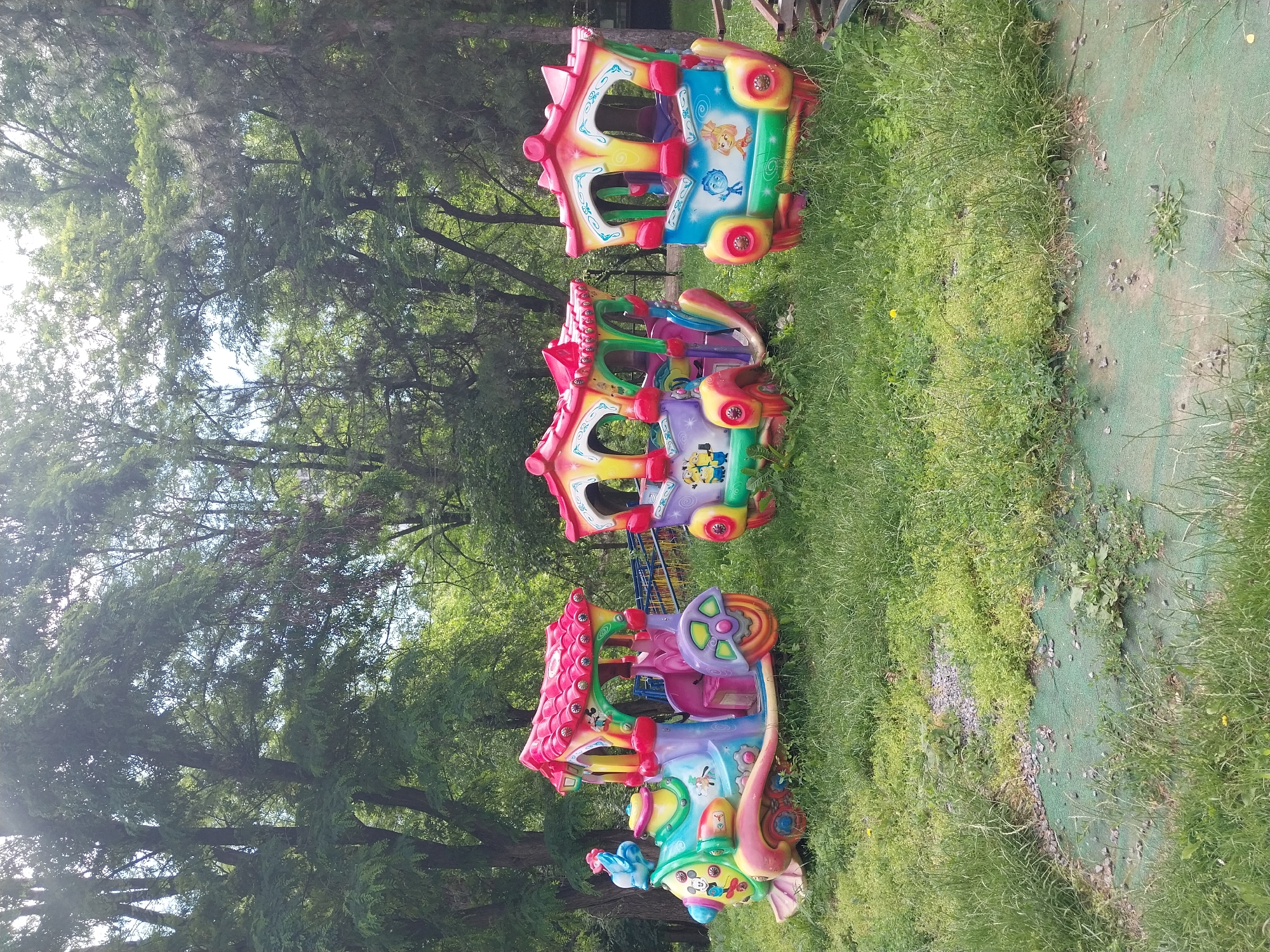 Three colorful, child-sized train cars are parked on a grassy area with a light green matting beneath. The train cars are decorated with cartoon characters, including Mickey Mouse and Minions. In the background, lush green trees create a park-like setting. The scene is likely in a city park or an amusement area within Dnipro, Ukraine, given the general aesthetic and the presence of children's rides. The lighting suggests it is daytime, with sunlight filtering through the trees. There are no people visible in the image.
