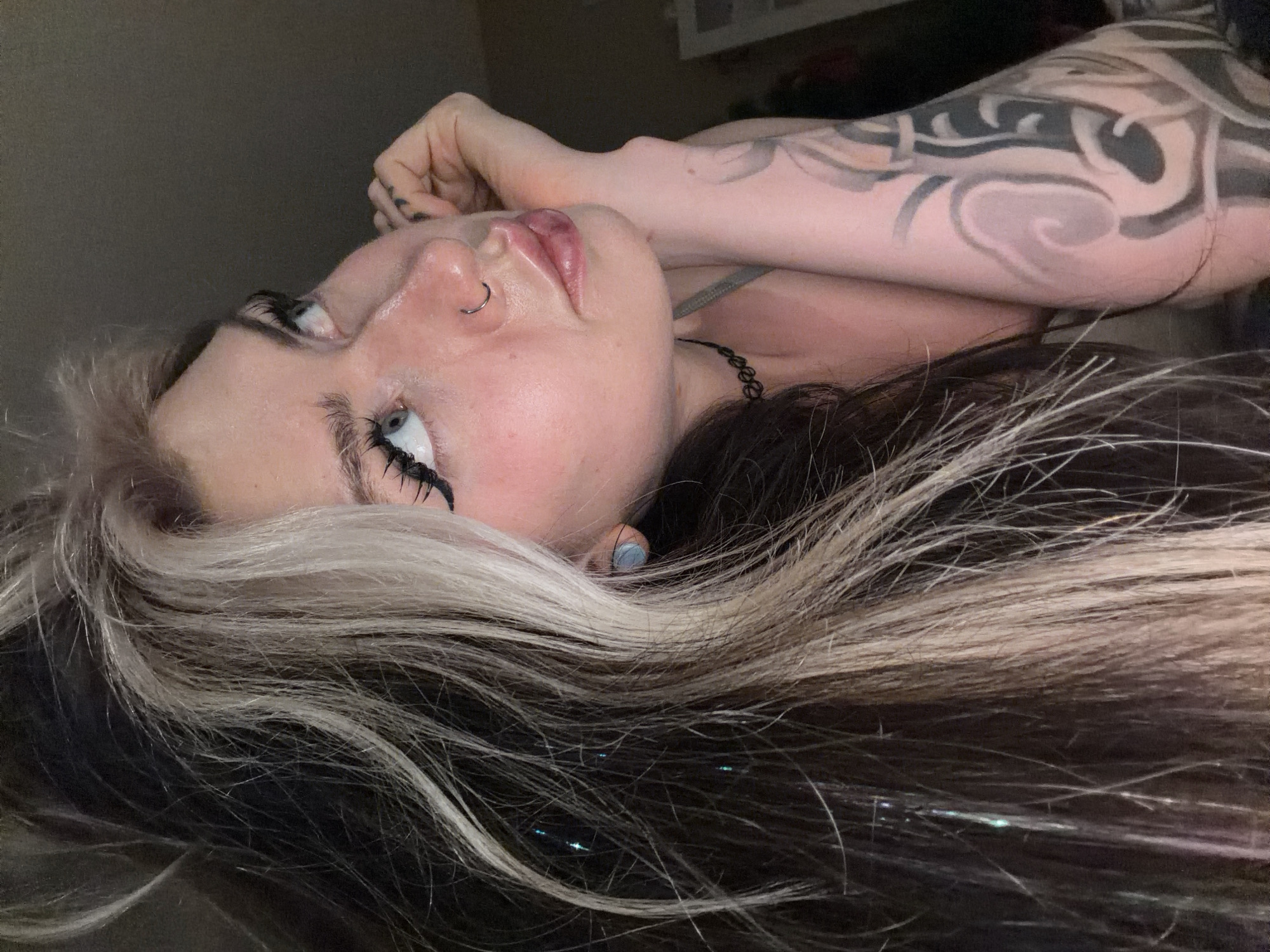 A woman lies on her back, her head angled towards the viewer. She has a fair complexion, blue eyes, and dark eyeliner. A silver nose ring is visible on her left nostril. Her hair is two-toned, with dark roots transitioning to platinum blonde highlights. Thin, metallic strands are interspersed throughout her hair. Her left arm is bent at the elbow and positioned behind her head, revealing a dark tribal-style tattoo on her forearm. She wears a dark choker necklace. The background is dimly lit and indistinct, suggesting an indoor setting.