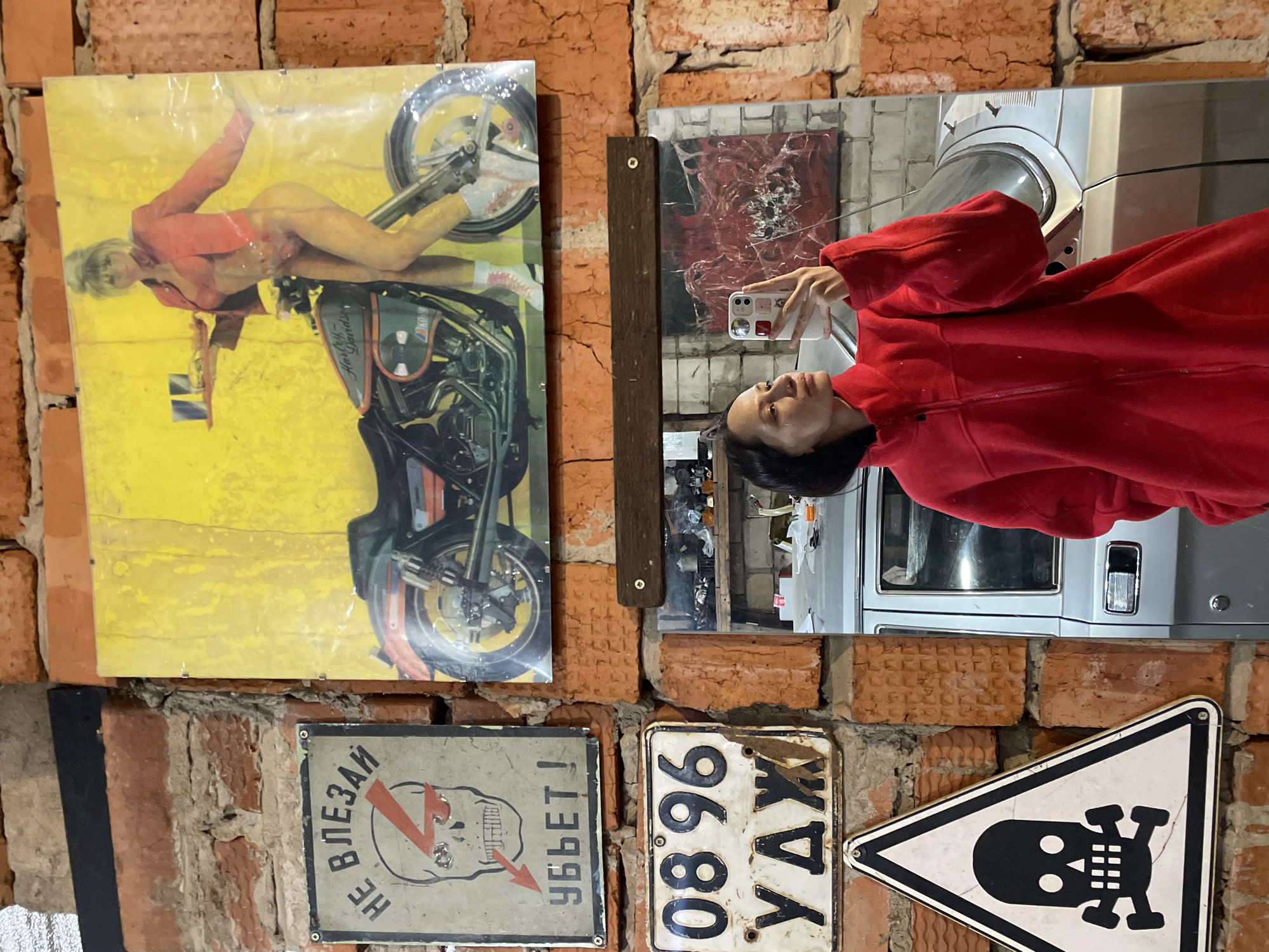 A person in a red hoodie takes a mirror selfie in front of a brick wall. The wall is decorated with several items, including a poster of a nude woman on a motorcycle, a bloody painting, a warning sign with a skull, and a license plate that reads "08 96 YAX." The reflection in the mirror reveals an interior space with a washing machine and other objects. The setting is identified as Oktyabr'skii, Russia.