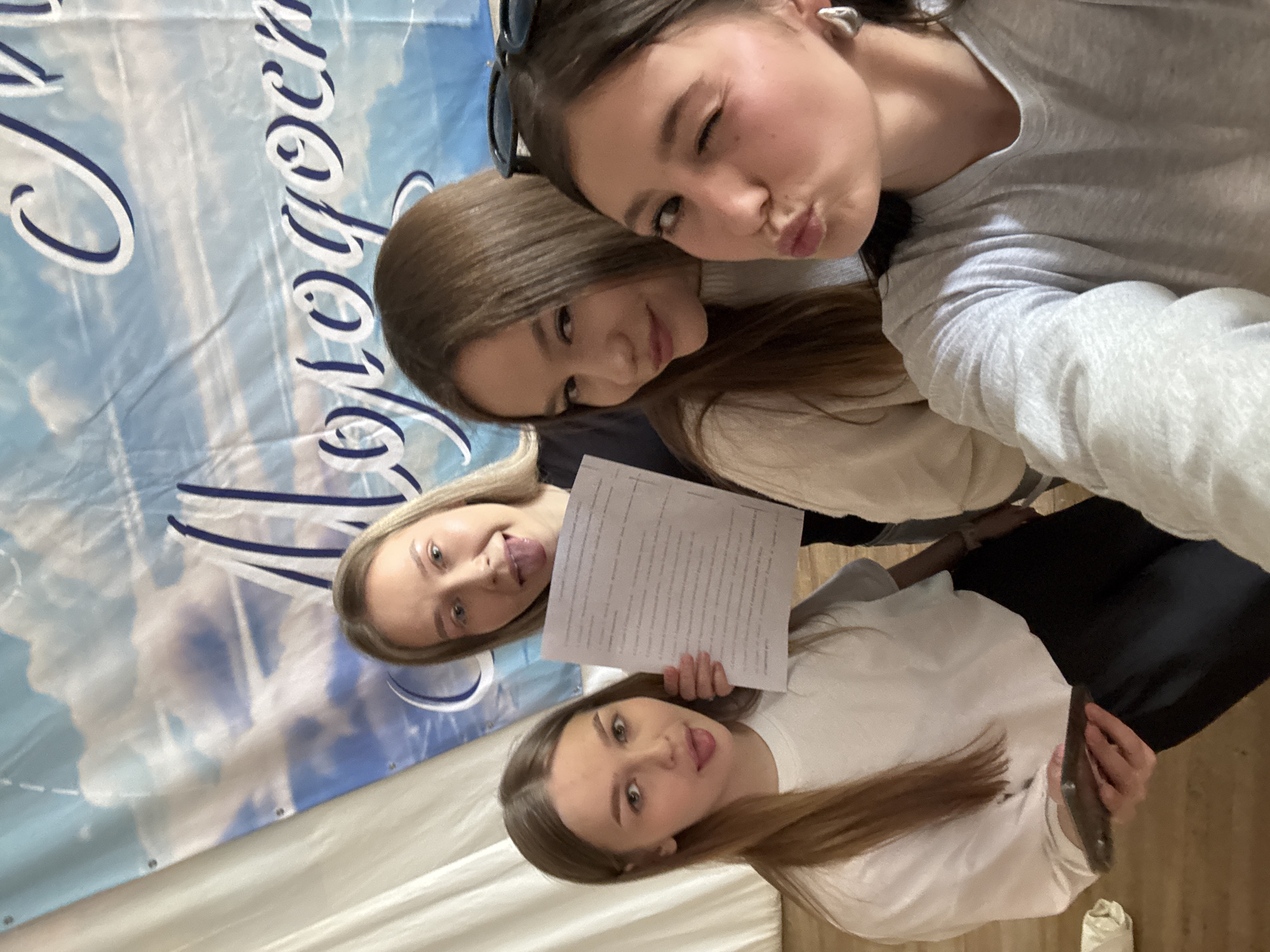 Four young women are posing for a selfie indoors. The background features a banner with blue sky and cloud imagery, with text in Russian. Two women are at the top of the frame, one winking and puckering her lips. Below them, another woman leans her head toward the top two. At the bottom, a fourth woman holds the phone. The location is the city of Chaikovsky, Russia. One woman holds a sheet of paper filled with text. The women are wearing casual clothing. The floor is a light wood laminate.