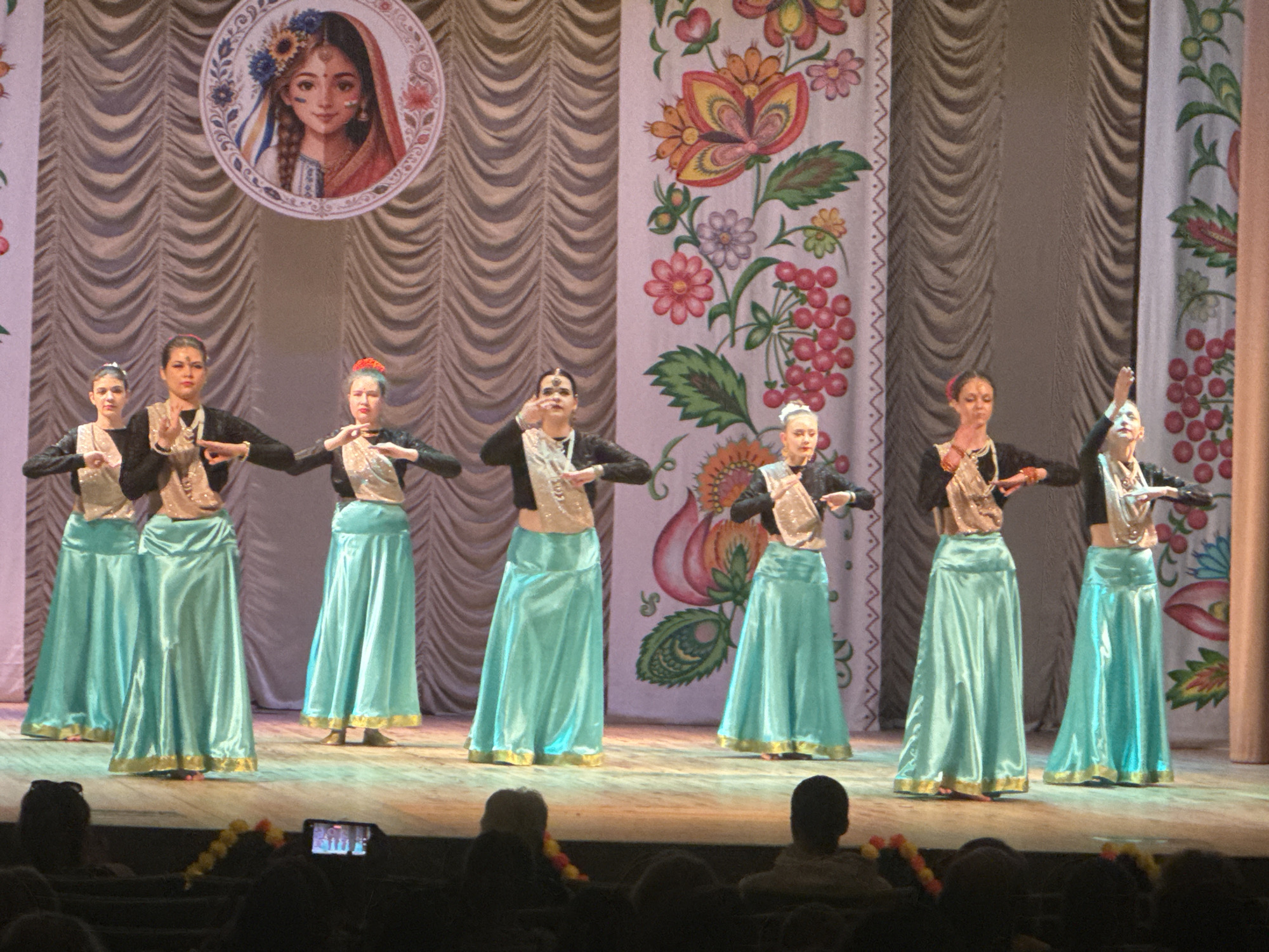 In an indoor performance space, five women in traditional Indian attire are standing on a stage, posing for a photograph. They are wearing teal-colored, floor-length skirts with gold trim, and black tops with embroidered vests and adornments around their necks. Each dancer has her hair pulled back and adorned with either a red or white flower. They are all making similar hand gestures, with their palms facing outwards. The backdrop of the stage features draped curtains and a large, circular image of a woman's face adorned with flowers and cultural symbols. The floor of the stage is illuminated by stage lighting, casting shadows and highlighting the dancers' costumes. In the foreground, out of focus, are audience members seated in darkened rows, with a cell phone recording the performance. The overall scene suggests a cultural dance performance or recital.