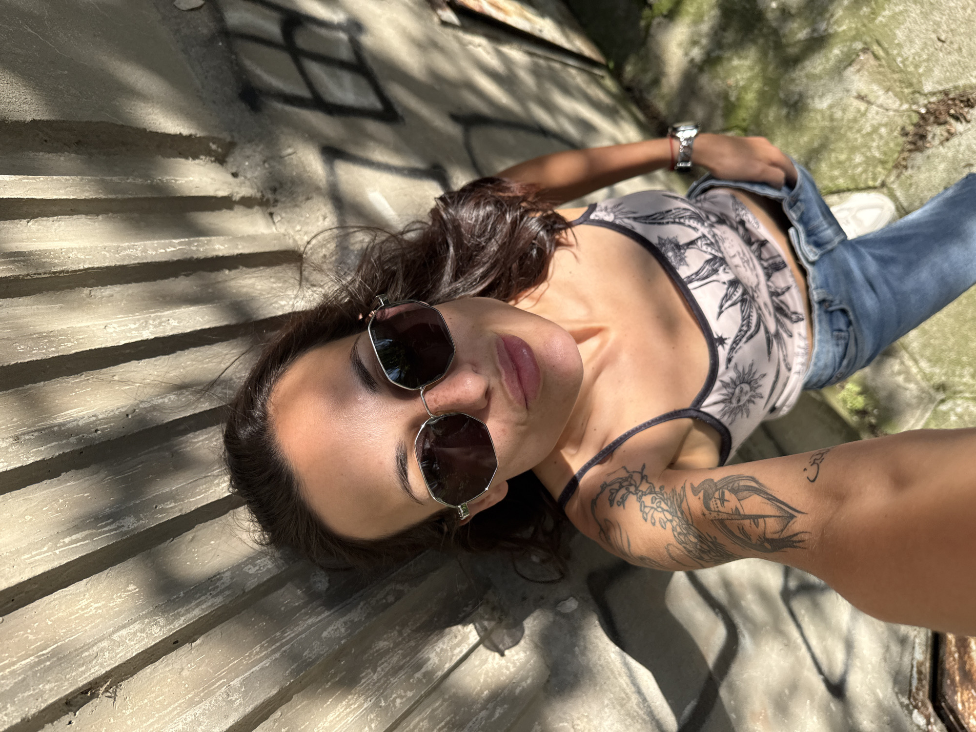 A young woman is lying on a set of concrete steps in what appears to be an outdoor setting. She is facing the camera and wearing sunglasses, a light-colored tank top with a dark pattern, and blue jeans. She has dark hair, and there are tattoos on her left arm. Her left hand is resting near her hip, and she is wearing a watch on her left wrist. The steps are light gray and show signs of wear and graffiti. The lighting is uneven, with shadows cast across the steps, suggesting a sunny day. Based on the information that the location is Pleven, Bulgaria, the scene could be within an urban area of the city, potentially in a park, near a building, or in an abandoned area where such steps are found.