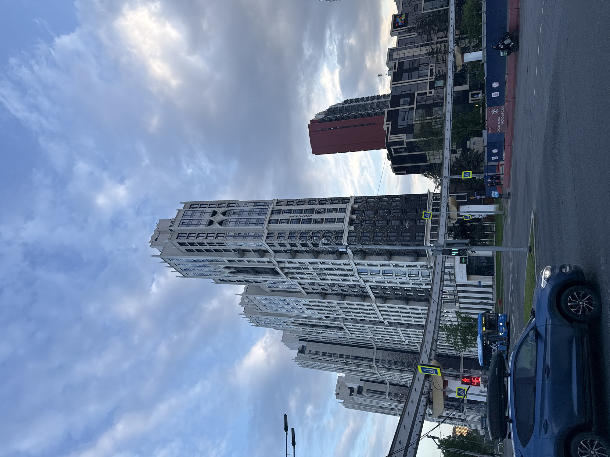 The image shows a street scene in Khimki, Russia, featuring a row of tall, modern residential buildings against a cloudy sky. The buildings are predominantly white and grey, with a mix of geometric patterns and multiple windows. A red building is visible further down the street.
In the foreground, a blue car is parked on the right side of the street, with its headlights on. A bus can also be seen further down the street. Streetlights and traffic signals are positioned along the sidewalk. A pedestrian crossing sign is also visible. A scooter is parked near a construction fence in the middle distance.
