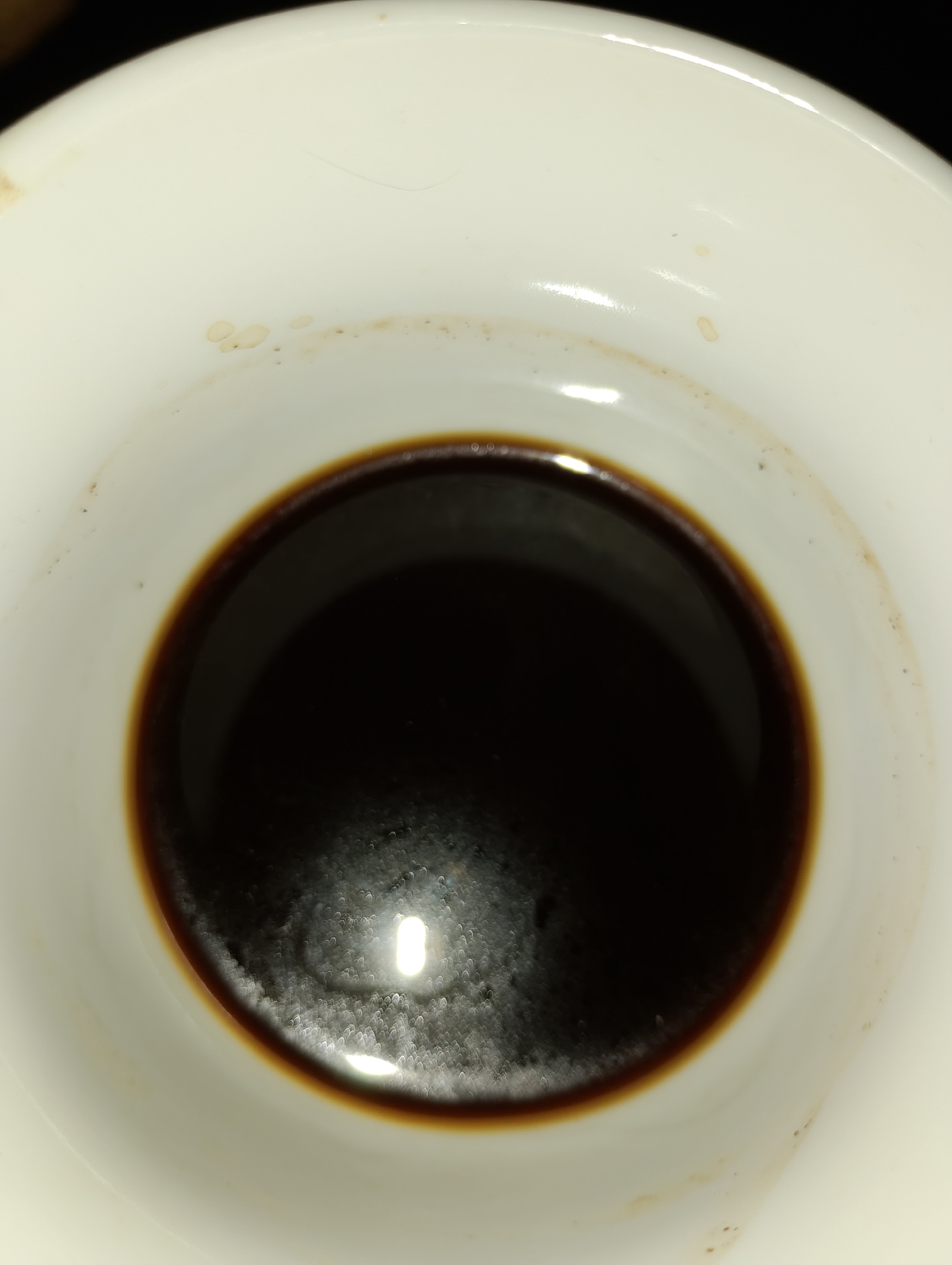 A close-up, top-down view shows the interior of a white ceramic cup filled with dark coffee. The coffee's surface is slightly foamy with small bubbles, reflecting light in scattered, oval shapes. The rim of the cup is visible, with a few small, light brown stains and one dark speck on the inner white surface. The coffee itself is a deep, dark brown, obscuring any view into the bottom of the cup. The lighting creates a strong reflection on the coffee's surface, making it difficult to discern fine details within the liquid.