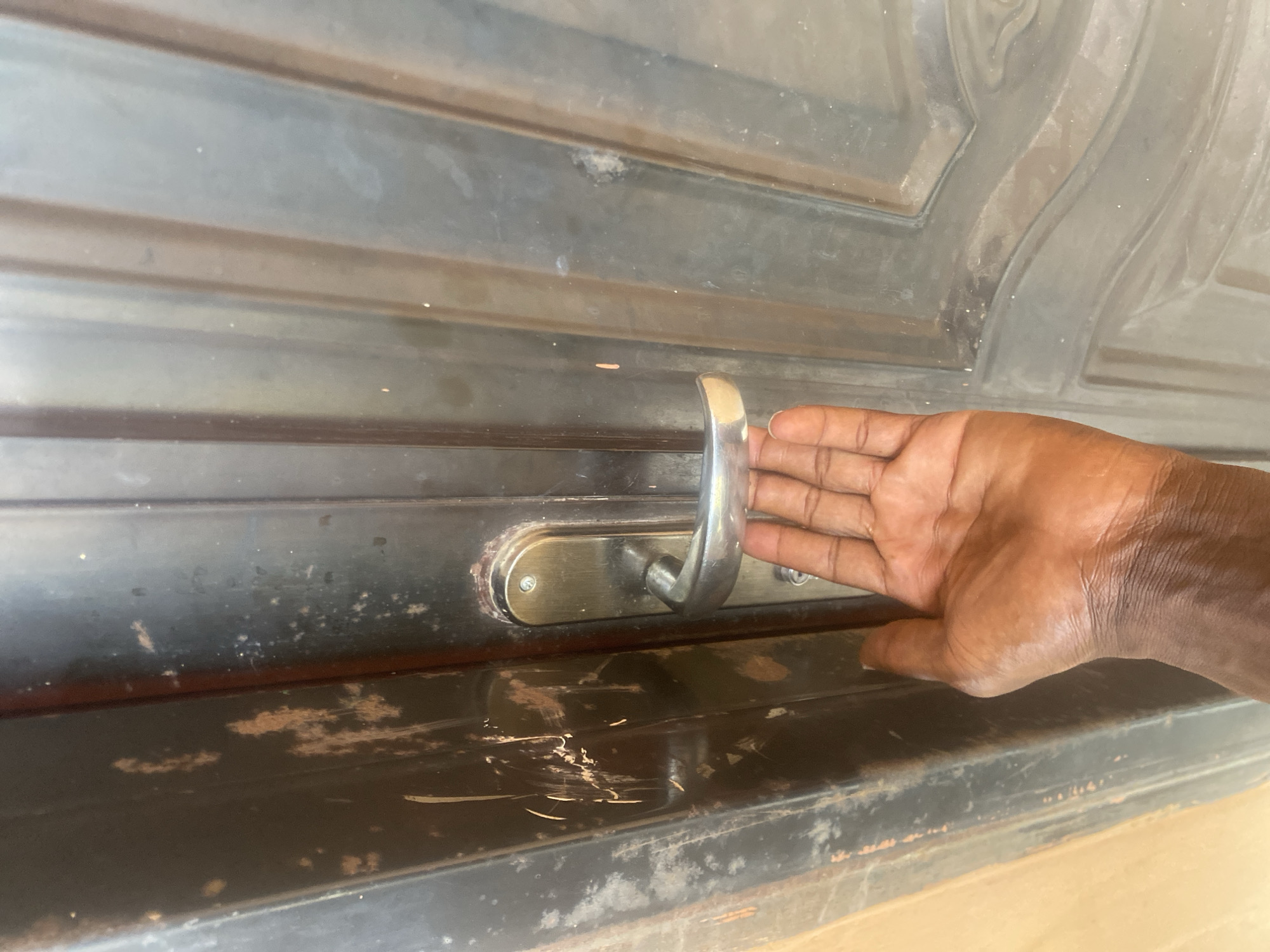 A close-up, eye-level shot shows a person's hand reaching for a silver door handle on a dark, textured metal door. The door appears worn, with visible scratches and rust spots, particularly on the left side. The hand is brown-skinned and positioned to grip the handle. The background consists of the door, which has vertical ridges and a patterned panel on the right side. The lighting is direct, casting subtle shadows. The overall impression is of an entrance to a building in Dumnga, Nigeria, suggesting a lived-in environment.