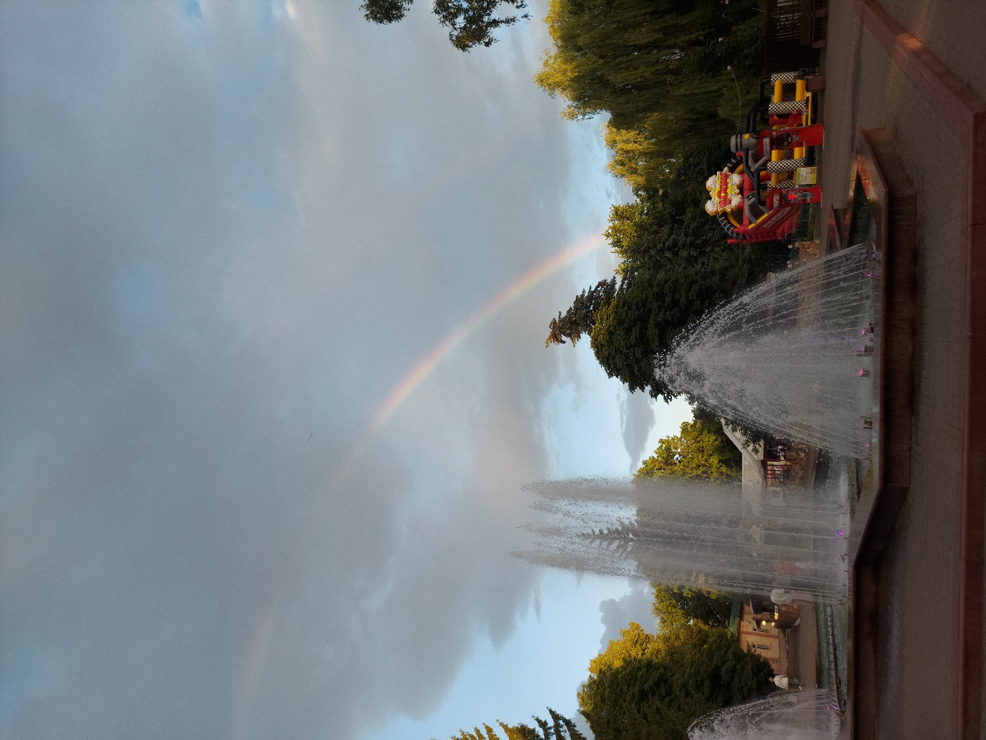 The image captures a scene at what appears to be an amusement park or fairground, possibly in Zheleznogorsk, Russia. The orientation of the image is portrait, meaning it's taller than it is wide. Dominating the left side of the frame is a dramatic sky filled with a mix of clouds and a visible rainbow arcing across it. The presence of the rainbow suggests it might have recently rained or there is ongoing light rain with sunshine.
In the background, on the right side of the image, several elements contribute to the setting. There are lush green trees, with some appearing to be deciduous and others possibly evergreens, framing the scene. Amongst the trees, there's a bright red and yellow amusement park ride with checkered accents. The ride has a whimsical design, with cartoonish elements and what looks like text on it, although it's not clearly legible. Further back, a building with a light-colored facade and windows is partially visible through the foliage.
In the foreground on the right, a large, multi-tiered fountain is spraying water high into the air, creating a misty effect. The water catches the light and appears dynamic. The fountain is situated on a paved area, possibly a walkway or plaza, with a reddish-brown edge. The overall impression is one of a festive atmosphere, possibly during a late afternoon or early evening, given the quality of the light and the dramatic sky. There are no clear individuals prominently featured in the image, though there might be some blurred figures in the distance near the building. The primary focus is on the natural phenomenon of the rainbow, the man-made attraction of the amusement ride, and the refreshing display of the fountain.