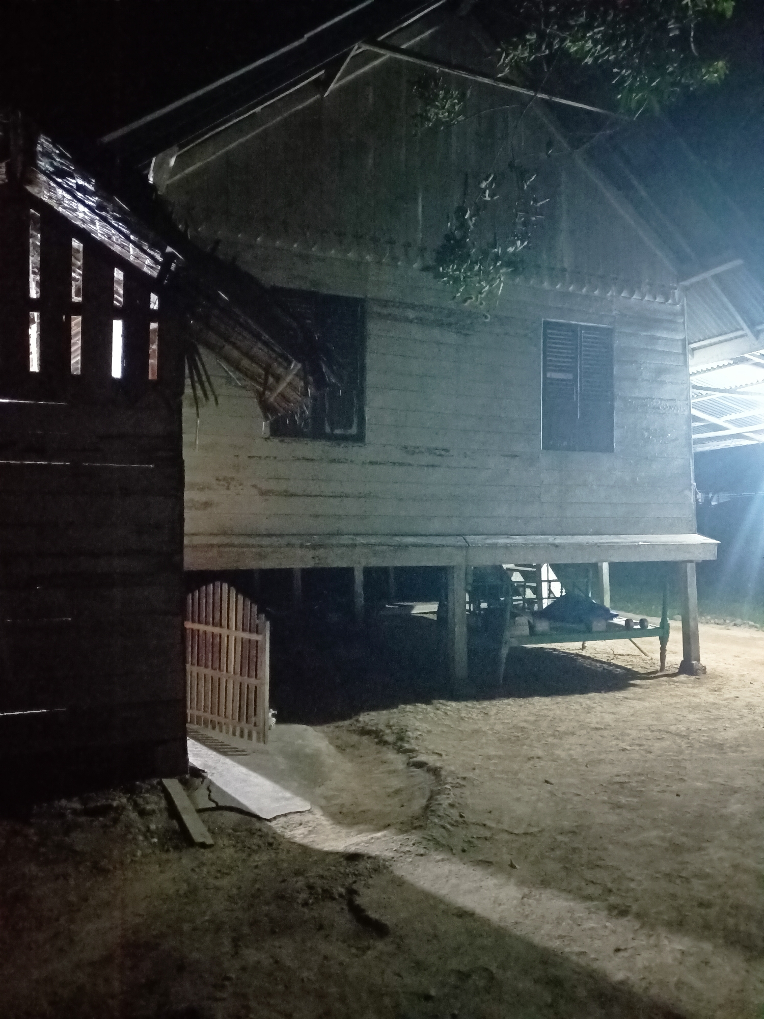 The image depicts an outdoor scene at night, likely in Asanbideun, Indonesia, featuring traditional stilt houses and surrounding ground. A stilt house, constructed from light-colored wooden planks, dominates the right side of the frame. It has a sloped roof covered with corrugated metal sheeting and two dark-colored windows with louvred shutters. The house is raised on green-painted stilts, with storage or objects visible beneath it, including what appears to be a green metal frame, possibly a bed or bench, with some items on it.
To the left of the stilt house is a darker structure, also wooden, with vertical slats that allow light to pass through from the interior, suggesting it might be another dwelling or a covered area. A small, light-colored wooden gate with vertical planks stands ajar between the two structures, leading to a pathway.
The ground is primarily dirt and sand, with patches of dry grass and a few scattered debris. Shadows are cast by the structures and a tree with foliage visible in the upper right, indicating artificial lighting from an unseen source. The overall atmosphere is dark and somewhat rustic. There are no people or animals visible in the image.