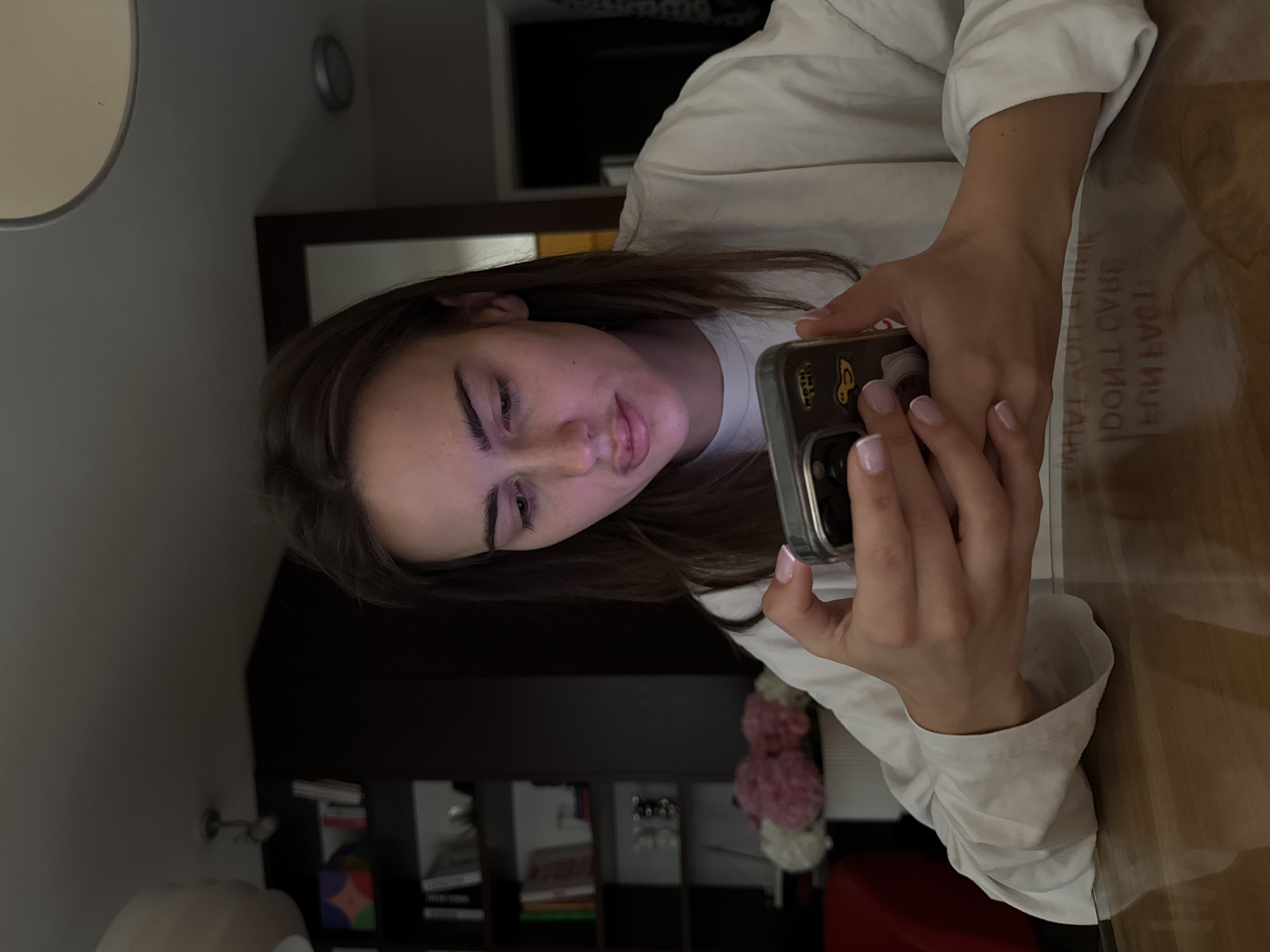 A young woman with long brown hair is seated at a table and looking at her phone. She is wearing a white shirt. The table has a wood-grain surface. Behind her, there is a bookshelf filled with books and a bouquet of flowers. The location is indoors, likely a home. Information about whether it is in the city of Warsaw, Poland cannot be determined from this image.