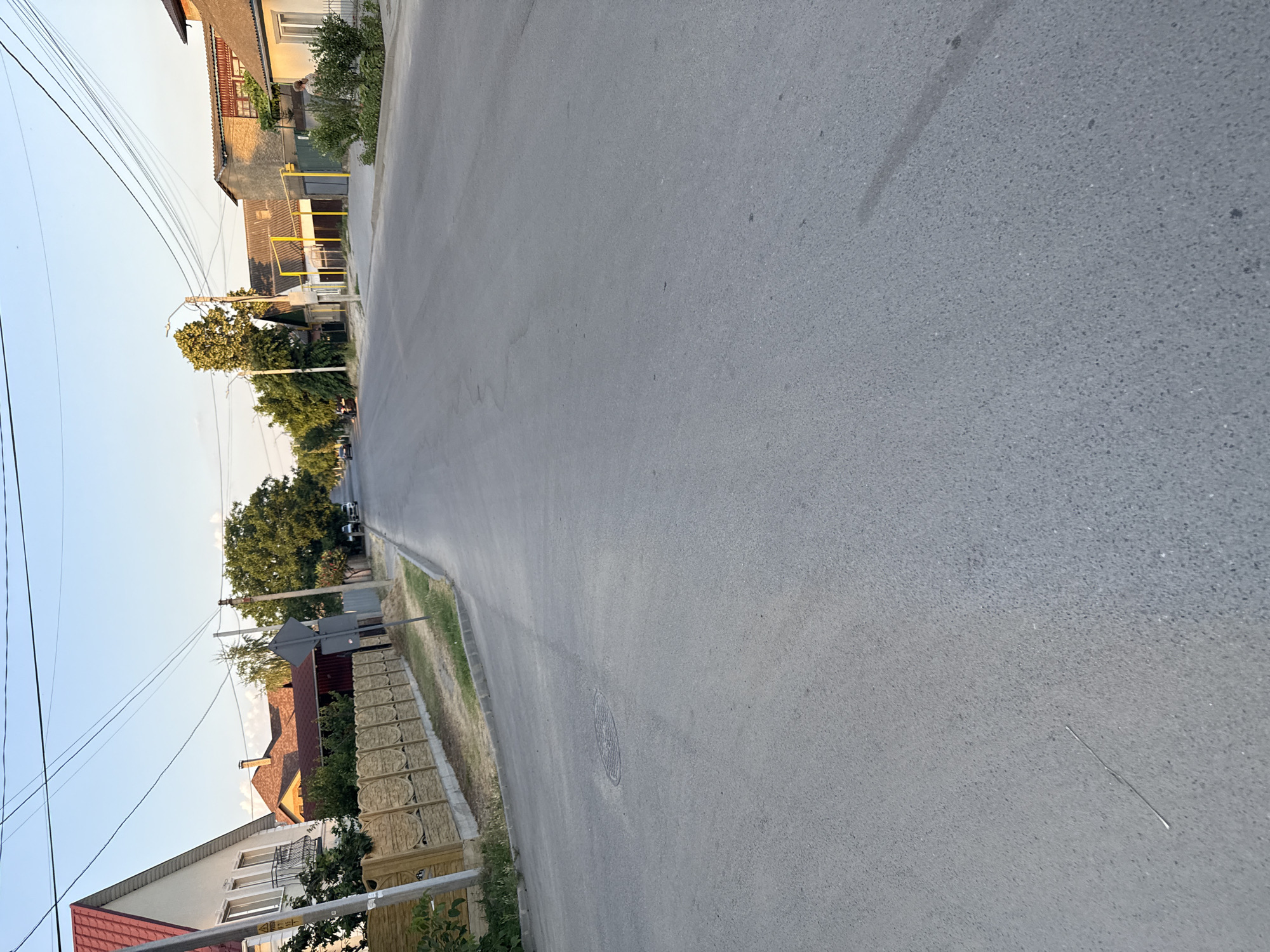 The image shows a street in a residential area. The road is paved and appears to be in good condition. On the left side of the road, there are houses and trees. The houses are of various sizes and styles, some with red tile roofs and others with lighter colored facades. There are power lines running along the street, supported by utility poles. A manhole cover is visible on the road. The scene is bathed in the warm light of what appears to be late afternoon or early evening, suggested by the long shadows. The sky is clear and blue, indicating fair weather. The environment appears to be a suburban or rural setting, possibly in Cahul, Moldova, as hinted by the location context. There are no people clearly visible in the frame, and the overall impression is one of a quiet, ordinary day on a residential street.