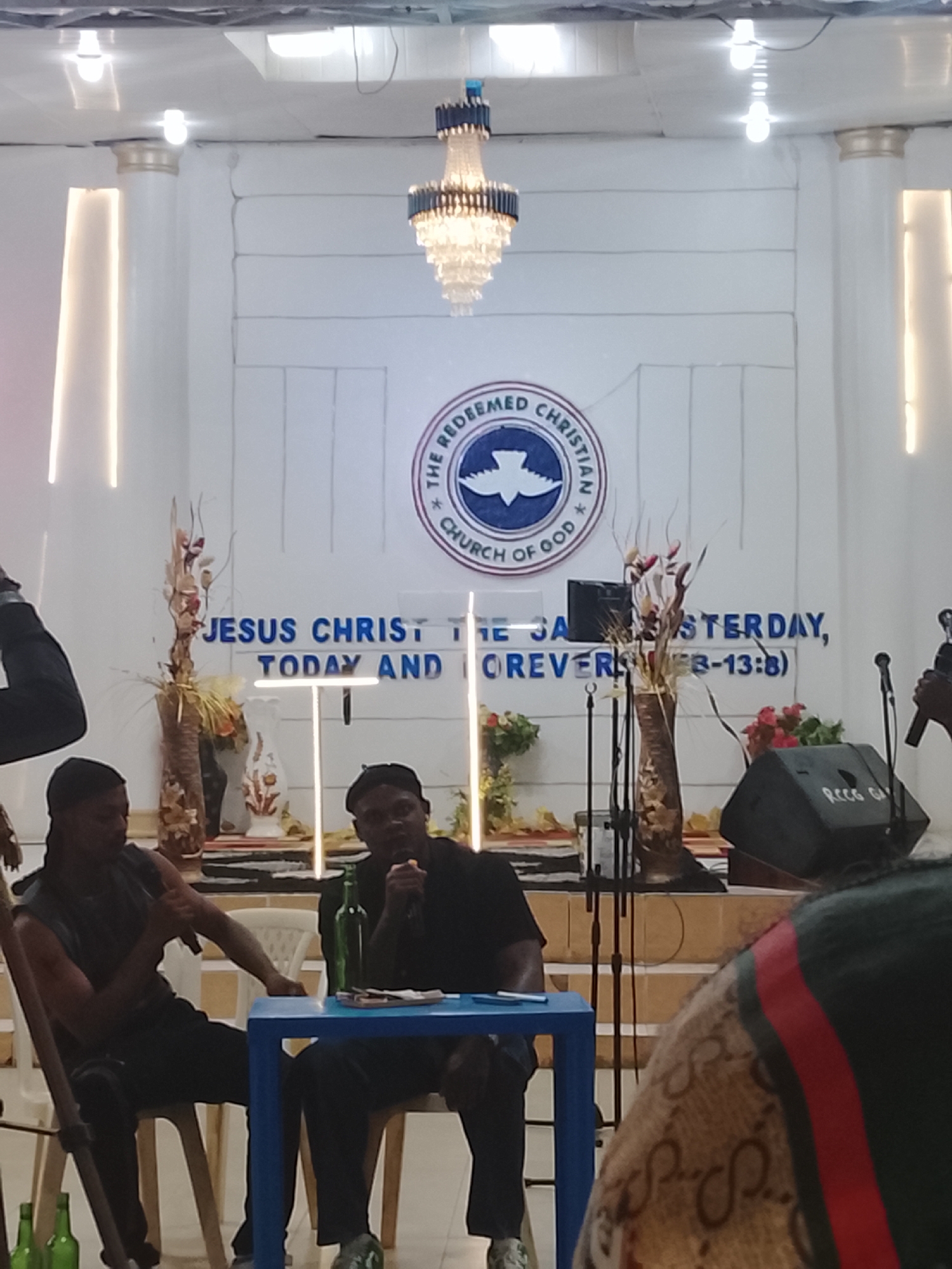 Here's a description of the image:
**Location:** The image appears to be taken inside a church auditorium. The church logo visible in the background is that of "The Redeemed Christian Church of God." There is not enough information to identify the location in Jos, Nigeria.
**People:** Two men are seated at a blue table. One wears a sleeveless top and the other a dark shirt. Both hold microphones and appear to be speaking. Parts of other people's bodies are visible on the edges of the image, indicating an audience or crowd.
**Objects:** A chandelier hangs from the ceiling. The church backdrop includes a logo and biblical text. A small blue table sits between the two men. There are bottles on the floor.
**Action:** The men are speaking into microphones, possibly engaged in a presentation, discussion, or performance.
**Environment:** The setting is formal, with a church stage and backdrop.