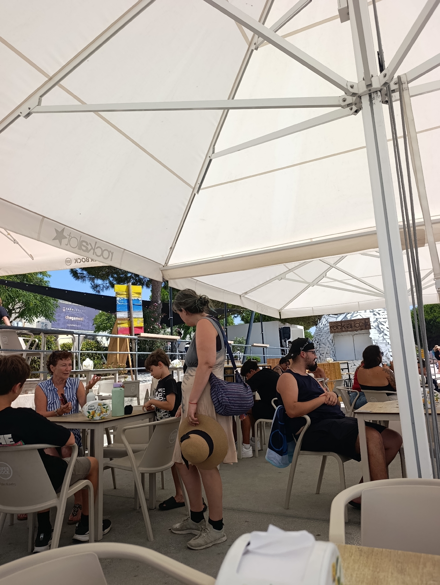 The image depicts an outdoor seating area under a large white canopy, likely part of a restaurant or cafe. In the foreground, a group of people are seated at tables. A woman with grey hair in a striped blue and white shirt is gesturing with her hands while talking to a young boy in a black t-shirt. A man wearing a black t-shirt and shorts is seated at another table, facing away from the camera. Further back, a man with a beard wearing a dark blue tank top and black shorts is seated, looking to his right. A woman with dark hair and a black top is also visible at a table. The environment appears to be sunny, with trees visible in the background. The presence of a sign with "TAGUSURBANIC" suggests a location in Setúbal, Portugal, likely within the TAGUSURBANIC area which is a vibrant cultural and leisure hub by the riverfront. The overall atmosphere suggests a casual outdoor gathering.