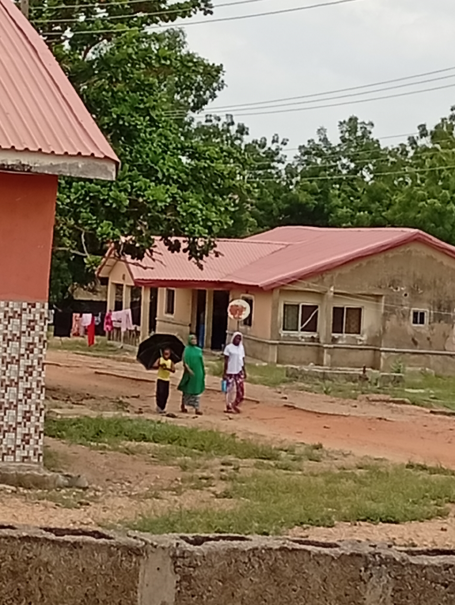 The image shows a dusty, rural scene in Adankolo, Nigeria. Three individuals are walking along a dirt path. A child, holding an umbrella, is followed by two women, one wearing a green headscarf and the other in a white shirt with a patterned skirt. In the background, there are buildings with reddish-brown roofs and drab exterior walls. Laundry hangs outside one of the buildings. The environment is characterized by trees and sparse vegetation. Overhead, power lines are visible. A concrete retaining wall is visible in the immediate foreground.