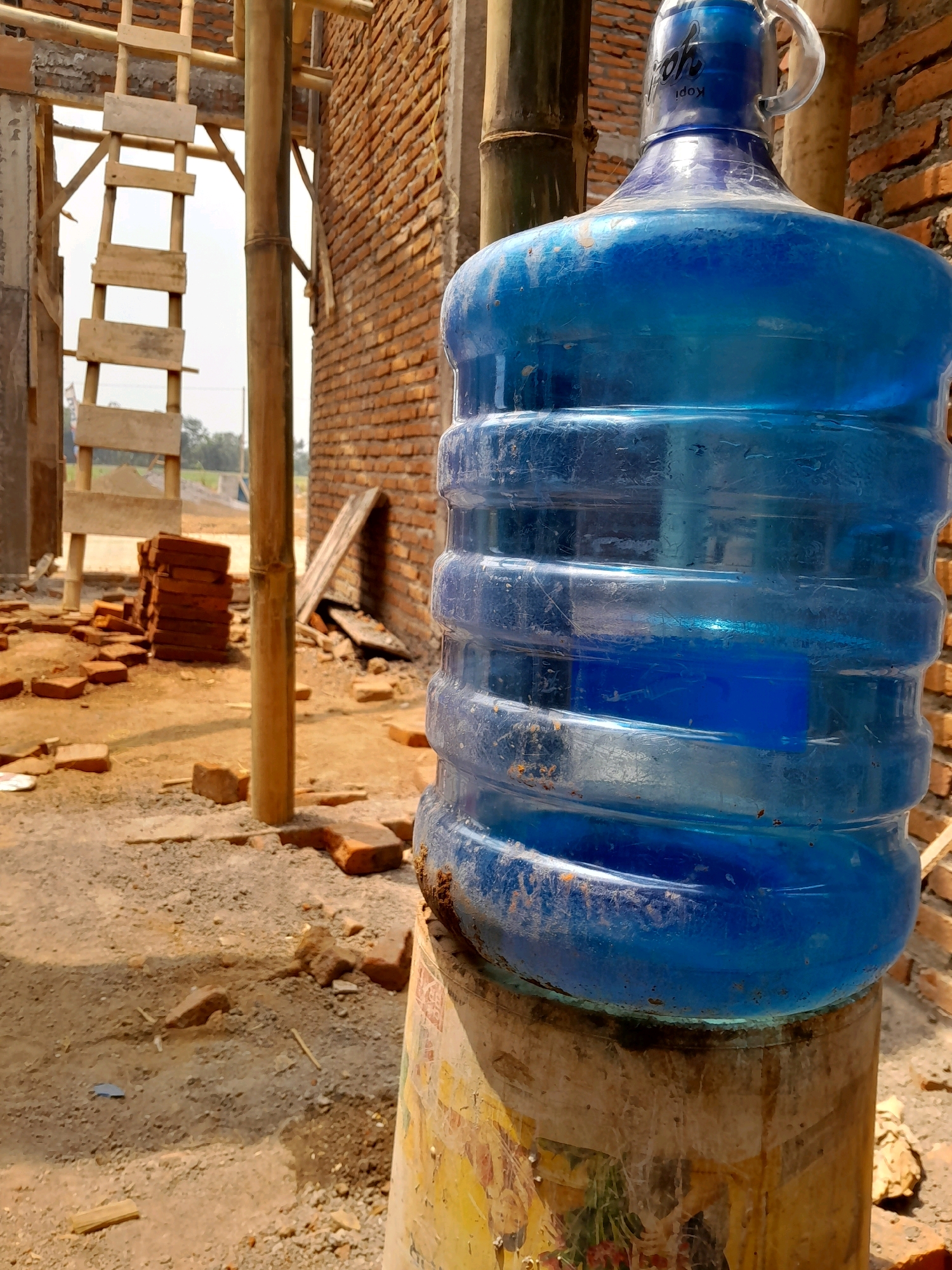 A close-up, low-angle shot depicts a large, translucent blue plastic water cooler jug positioned on top of a worn, cylindrical base, likely a dispenser, with faded colorful graphics. The jug is filled with water and shows numerous scuffs and dirt marks, particularly near its base. It has a clear plastic handle and a blue cap with white lettering that reads "Kopi Yooh."
The background suggests a construction site in Karangduren, Indonesia. A rough brick wall, built with reddish-orange bricks, forms the right side of the scene. To the left, wooden scaffolding and a ladder made of rough-hewn planks are visible, supported by bamboo poles. The ground is uneven and covered in dirt, gravel, and scattered red bricks, some broken. In the mid-ground, a stack of neatly piled red bricks sits on the ground. Further back, out of focus, is a blurry landscape with greenery and a bright sky. The lighting indicates it is daytime, with strong sunlight casting shadows.