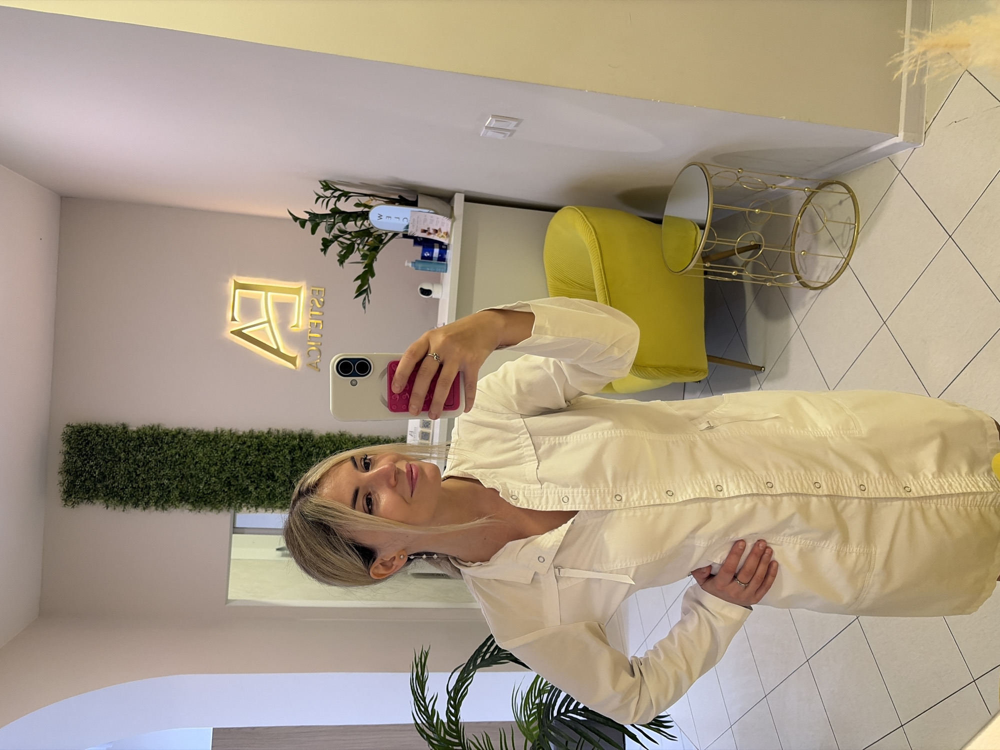 A woman in a white lab coat takes a mirror selfie in what appears to be a beauty salon. She has blonde hair pulled back in a ponytail and is wearing earrings and a ring. In the background, there is a green wall, a bright yellow chair, a small round table with a mirrored top, and a shelf with various beauty products. A neon sign with the letters "EA" and "ESTETICA" is visible on the wall. The salon appears to be clean and modern, with tiled floors. The woman is holding a phone with a pink case and is looking at her reflection. The overall impression is one of professionalism and contemporary style.