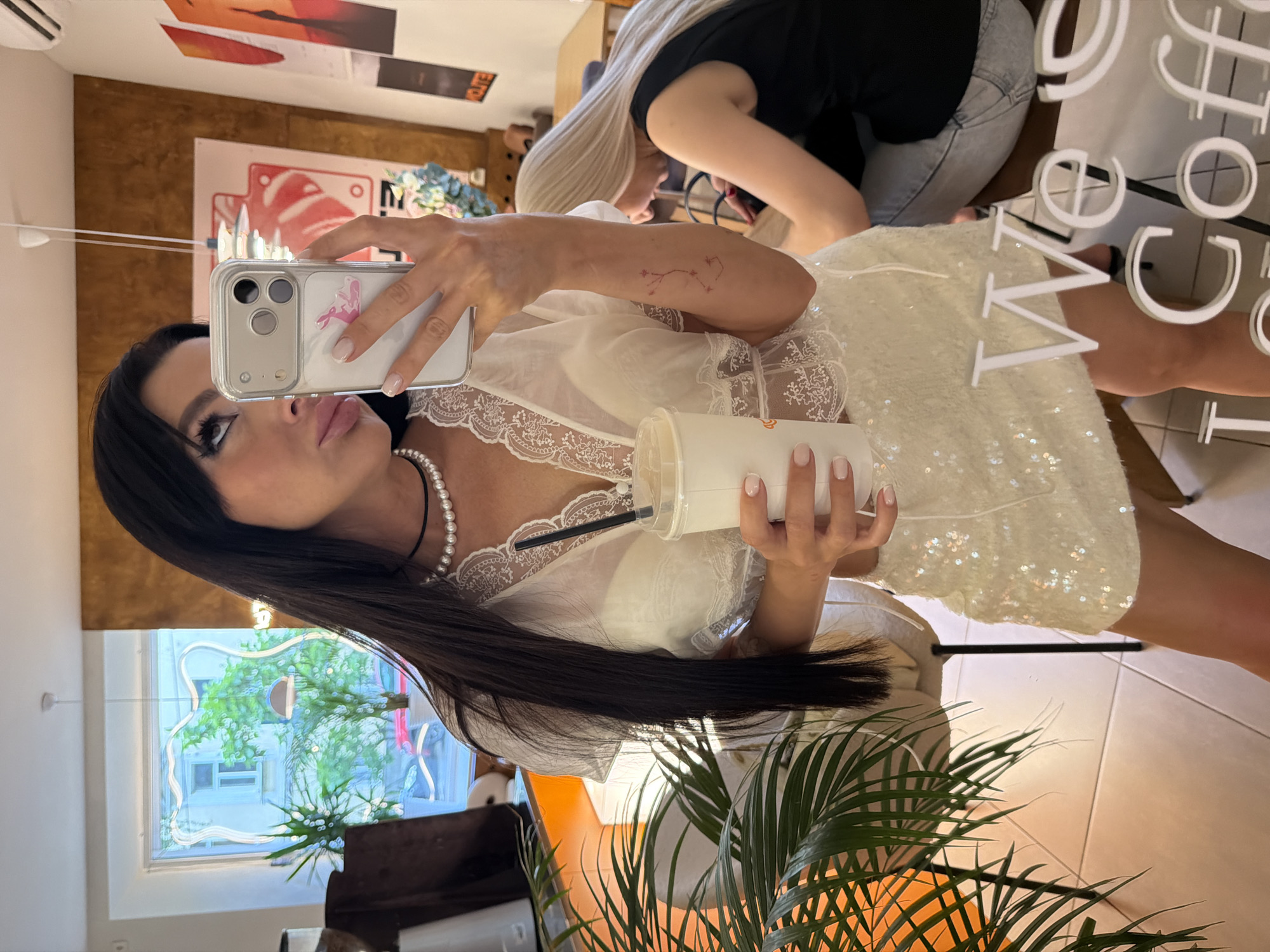A woman with long dark hair and heavy eye makeup takes a mirror selfie in what appears to be a cafe in Novaya Usman', Russia. She is wearing a white sequined skirt and a sheer white top with lace trim, and is holding a white paper cup with a straw in her right hand. She is wearing a pearl necklace. Her left arm is visible and has a small tattoo of constellations. Another woman with blonde hair is visible in the background, seated at a table. The cafe has a window with a view of trees and buildings, and posters on the wall. A large plant is in the foreground on the left. A mirrored surface with the text "We Coffee" is visible at the bottom of the image.
