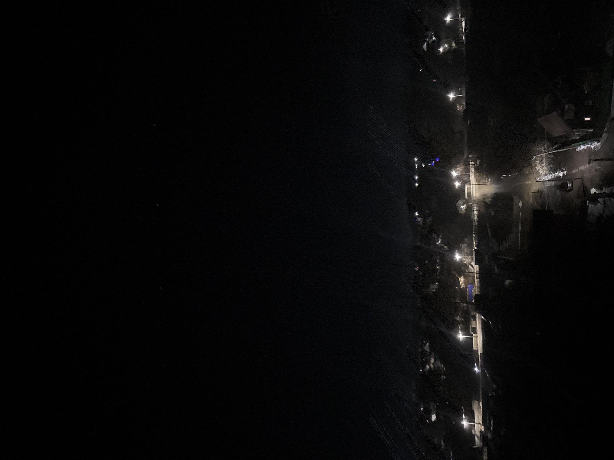 The image shows a nighttime view of Chaikovsky, Russia. Street lights illuminate a road running along a body of water. The left side of the image is dominated by the dark water, with reflections of some lights visible on the surface. The right side shows a road with streetlights and buildings alongside it. Some residential structures and possibly parked cars are discernible. Vegetation is seen along the road. The image is dimly lit, with most details obscured by darkness.