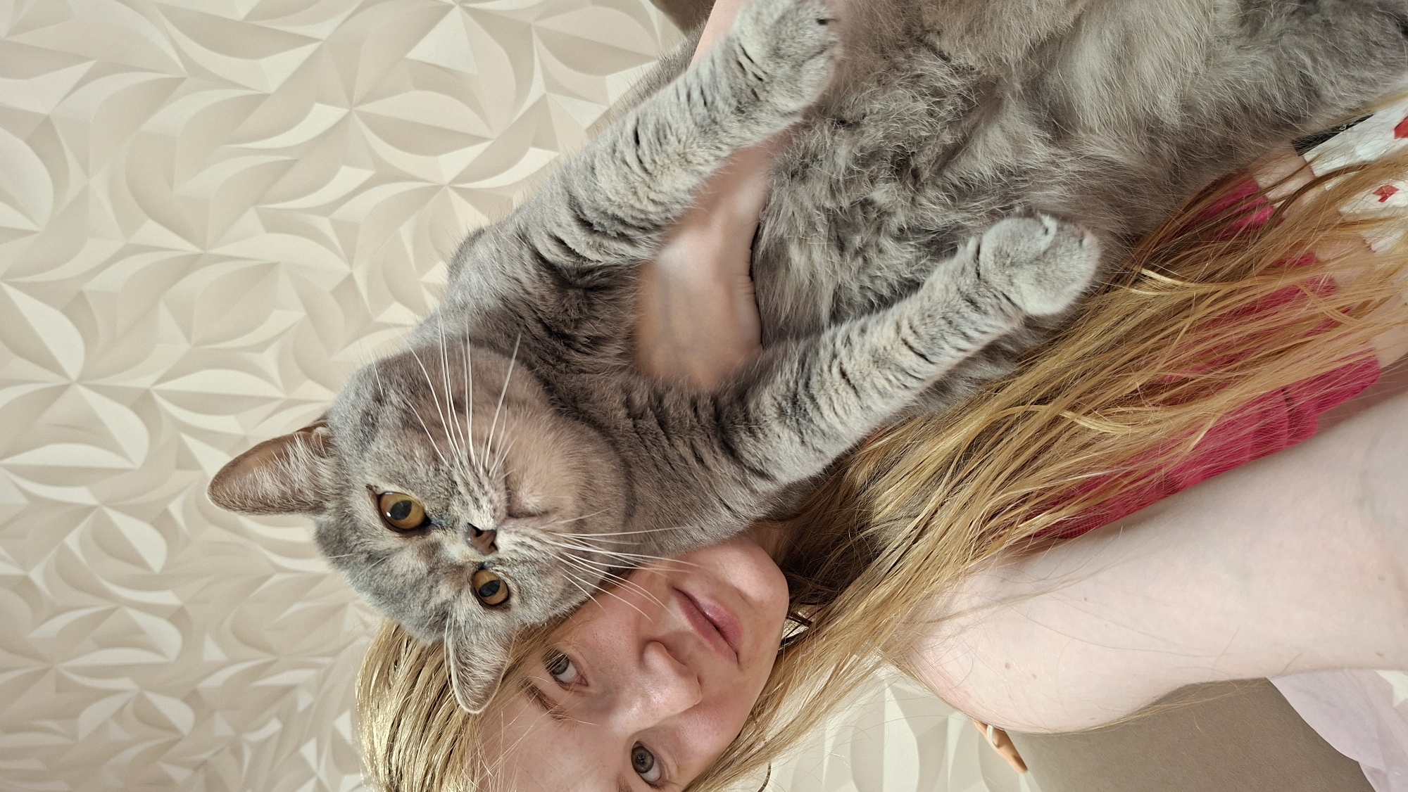 A young woman with long, blonde hair and fair skin is holding a grey, fluffy cat. The woman is wearing a pink top. She is looking towards the camera with a slight smile. The cat is looking away from the camera, towards the upper right. The background is a textured, light-colored wallpaper with a repeating geometric pattern. The scene appears to be indoors, possibly in a living room. The location is not specified in the image.