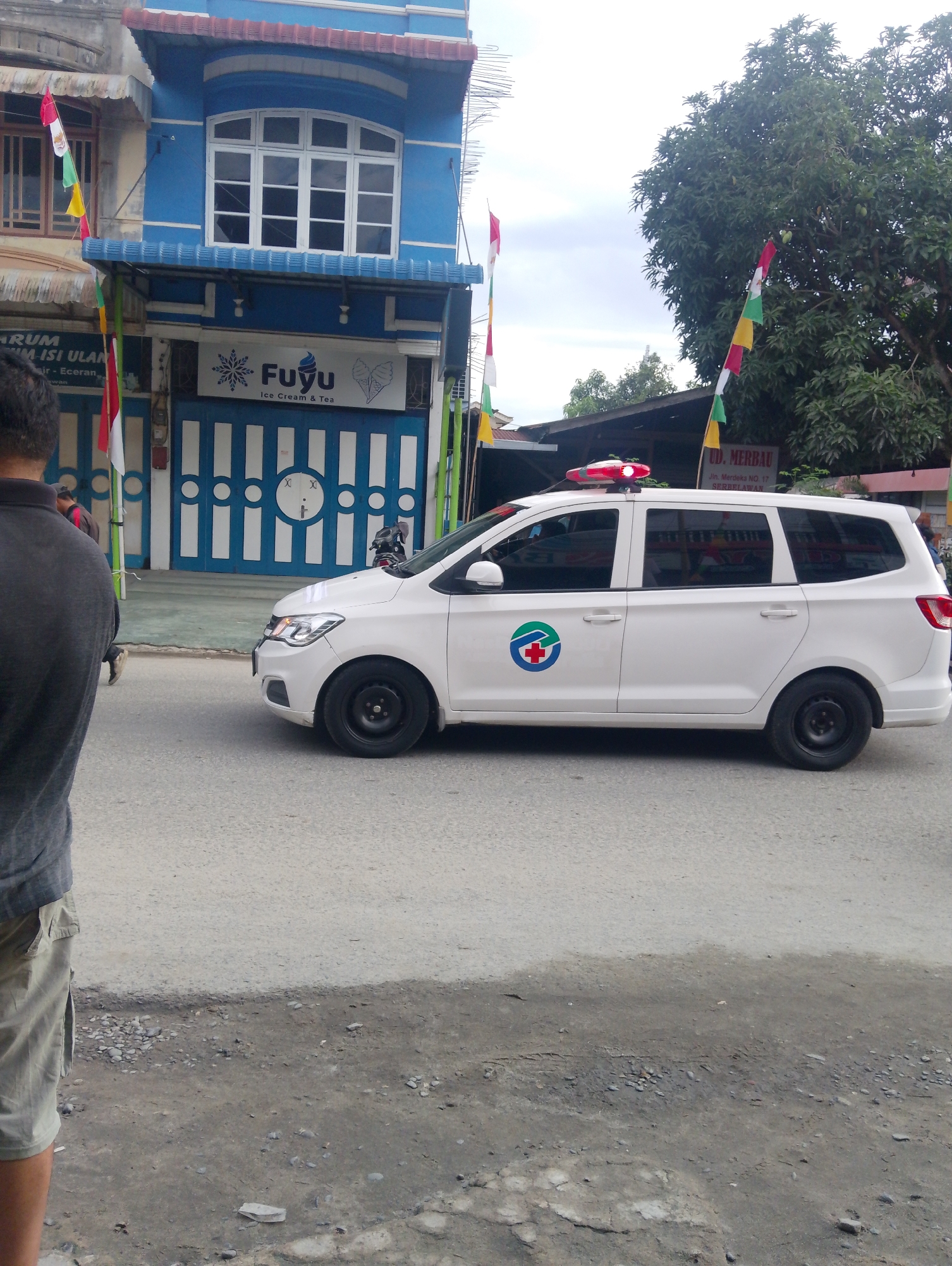 In Serbelawan, Indonesia, a white ambulance with red and blue markings, including a red cross symbol, is parked on a paved road. The ambulance has a red flashing light on its roof. To the left of the ambulance is a blue and white building with multiple windows and an awning. Flags are visible on the building and in the surrounding area. A person wearing a gray shirt and khaki shorts stands on the left side of the frame, facing away from the camera. In the background, a large green tree with some fruit is visible, as well as other buildings and vegetation. The ground is a mix of pavement and dirt.