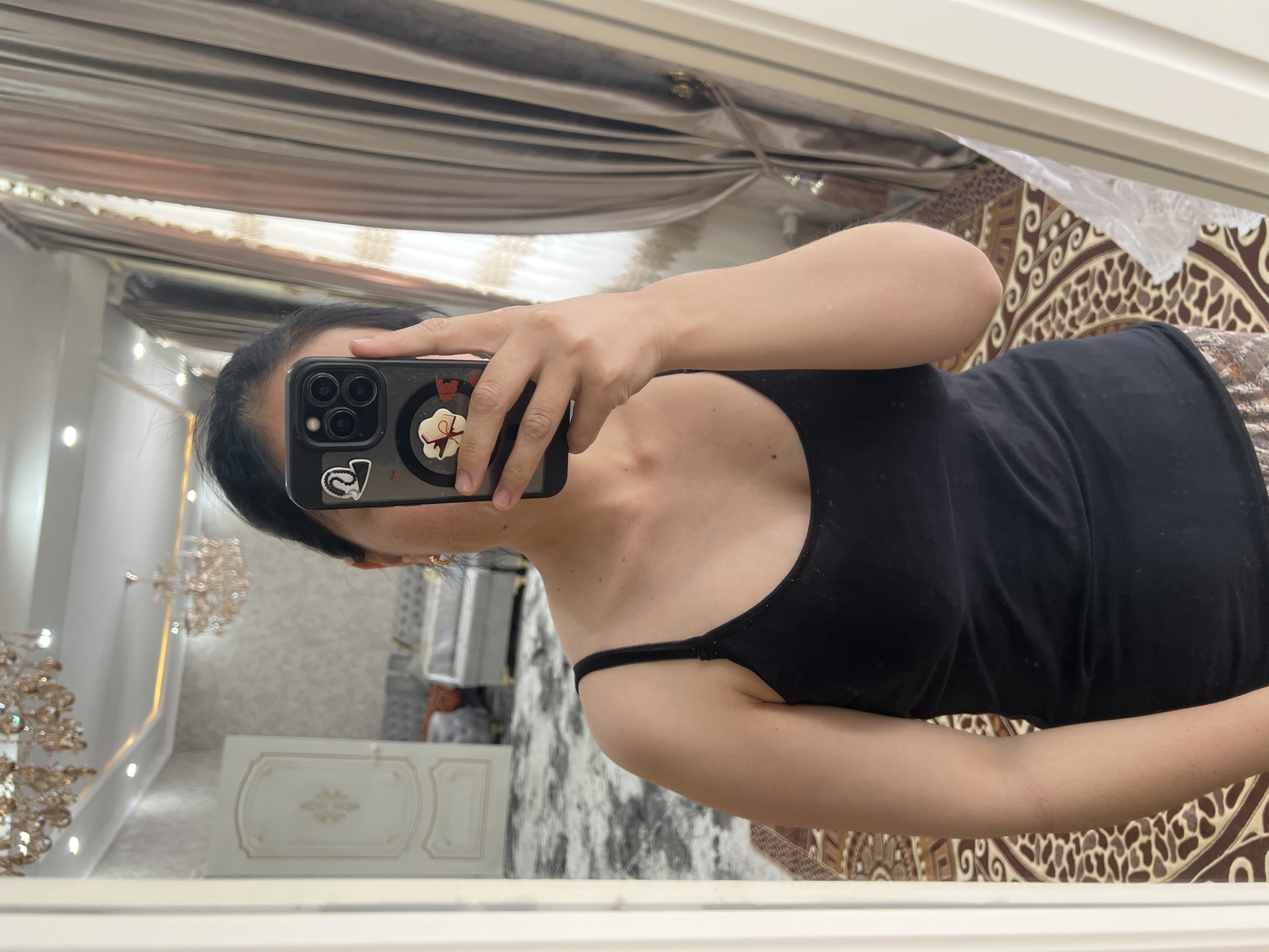 The image shows a woman taking a mirror selfie. She is wearing a black tank top and patterned pants. Her phone, with stickers on the case, is held up to obscure her face. In the background, a luxurious room is visible, featuring a chandelier, ornate wallpaper, a door, and draped curtains. The floor is covered by a patterned rug. The location is identified as Kara-Dıykan, Kyrgyzstan.
