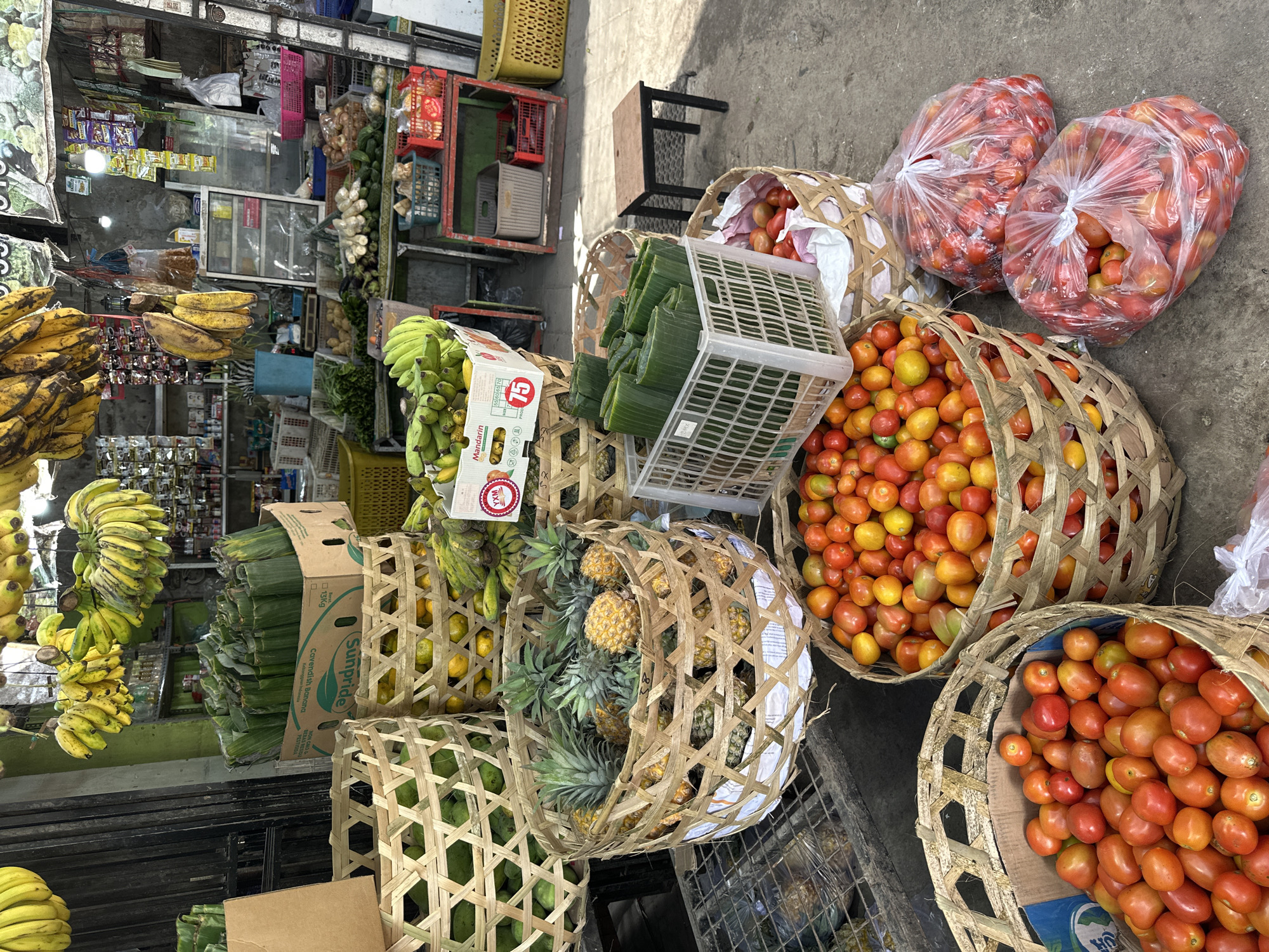 This is a photograph of a fruit and vegetable stand at a street market in Kuta Selatan, Indonesia. The scene is outdoors on a paved surface. In the foreground, there are several large woven baskets filled with produce. The largest basket on the left contains green mangoes. To the right of that, a basket is filled with oranges. Another basket in the middle is overflowing with pineapples. In front of the pineapples, a white plastic crate holds bundles wrapped in green leaves, likely vegetables. To the right of the crate, a smaller basket is also filled with pineapples. In the lower right foreground, two large woven baskets are filled with tomatoes, and two transparent plastic bags full of tomatoes are placed on the ground. In the background, shelves are stocked with various packaged goods, and bunches of bananas hang from the ceiling. A small wooden stool sits to the right of the produce display.