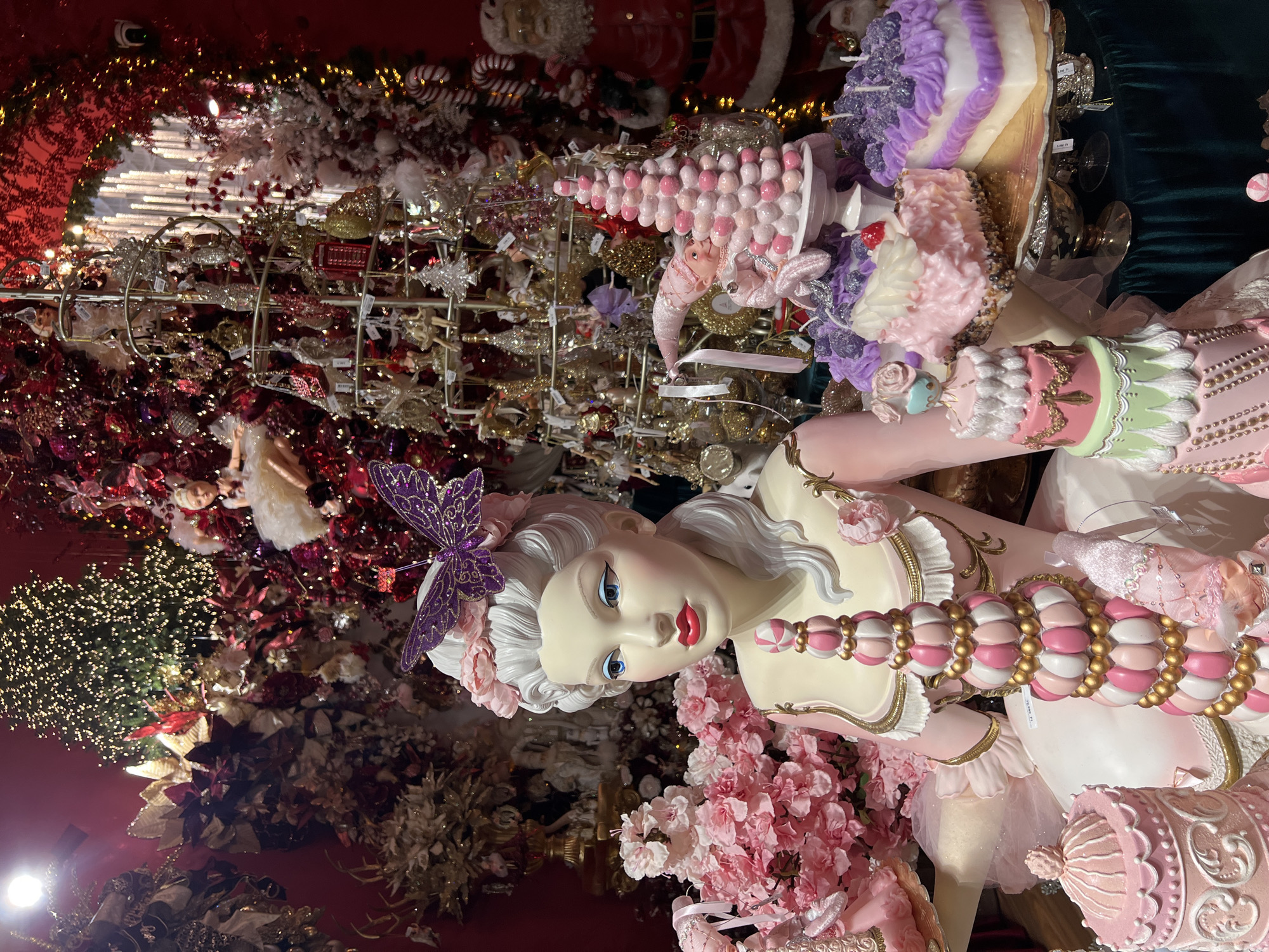 This image depicts a detailed and ornate shop window display in Budapest, Hungary. The centerpiece is a life-size mannequin made to look like Marie Antoinette, positioned among a vast array of sweets-themed Christmas decorations. The mannequin wears a pink dress, has a purple butterfly in her white, coiffed hair, and holds a large, pink and gold candy-like object.
Behind the mannequin are numerous decorations including cake-shaped ornaments, pink meringue-style Christmas trees, and countless gold, silver, and pink ornaments hung on metal stands. The backdrop consists of red and gold floral arrangements interspersed with strings of Christmas lights, as well as a Santa Claus. The overall aesthetic of the display is a mix of rococo elegance and whimsical sweetness.