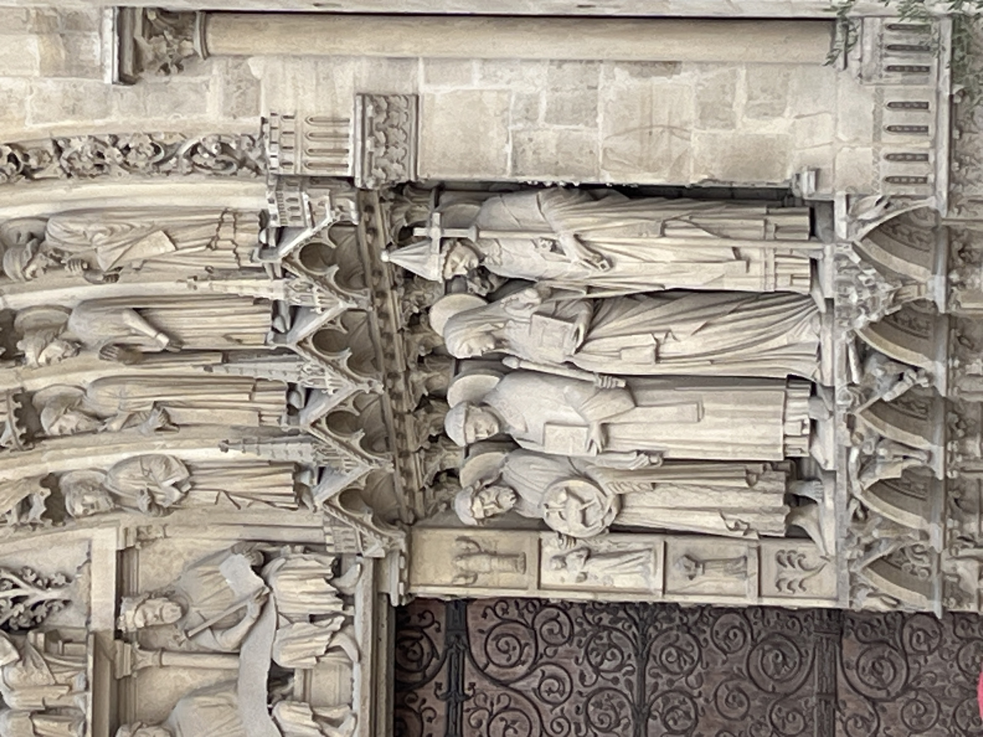 The image shows a close-up view of the sculptures on the facade of Notre Dame Cathedral in Paris, France. The sculptures depict several figures in religious attire, carved in stone. From left to right, there are four figures in the foreground standing on a ledge. The first figure holds a circular object with a lamb on it. The second figure holds a book. The third figure, a woman with a veil, also holds a book. The fourth figure, a man wearing a tall pointed hat and holding a cross, appears to be a bishop. Above these figures, other sculptures are visible, including a seated figure with a crown and scepter, and several standing figures in robes. The architecture features ornate carvings and arched details. The overall scene is one of religious and historical significance, characteristic of Gothic architecture.