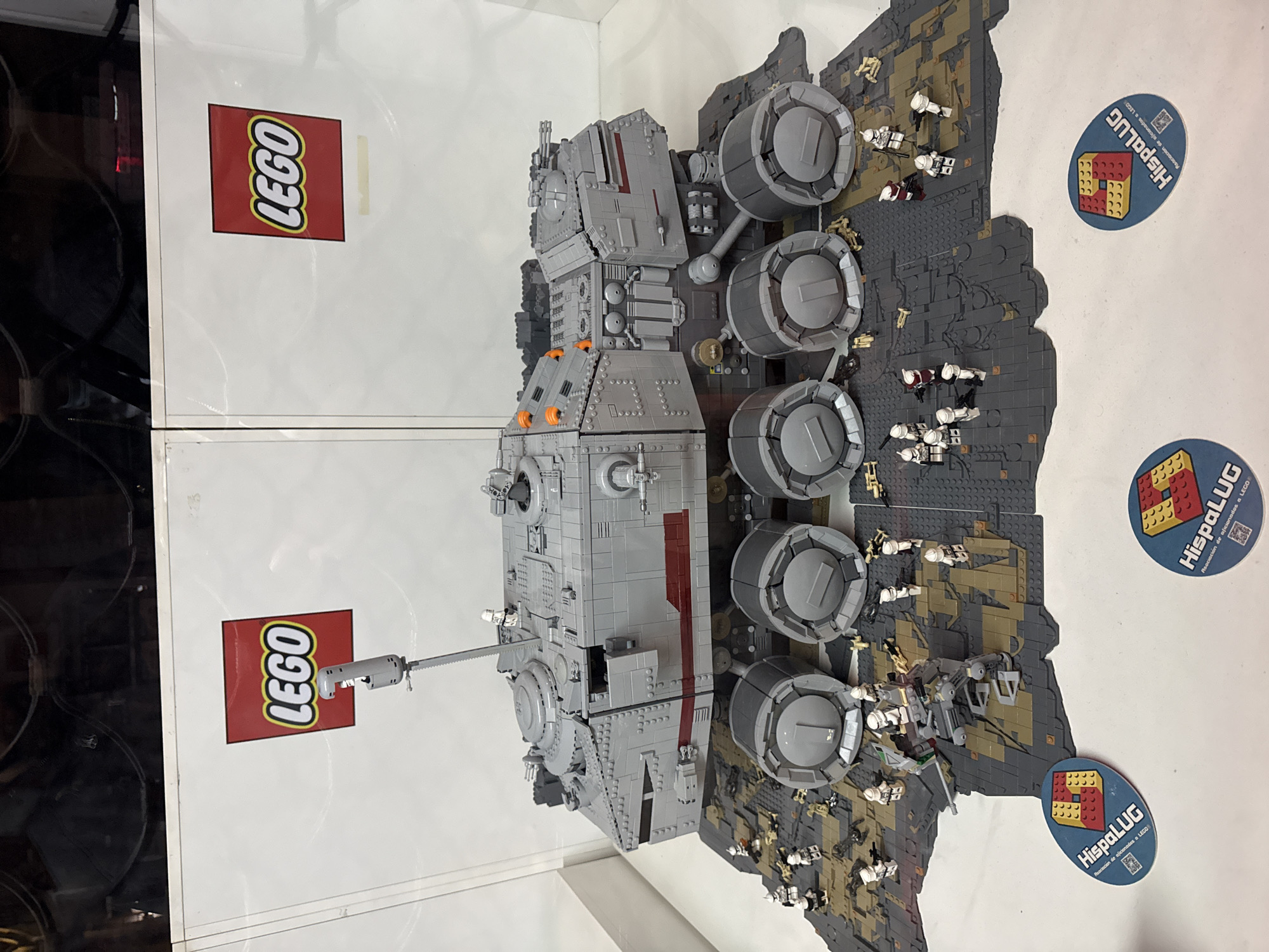 The image shows a Lego model of a Republic All Terrain Tactical Enforcer (AT-TE) from the Star Wars universe. It is displayed on a gray terrain with many Lego clone trooper minifigures around the AT-TE. There are stickers with the text "HispaLUG Asociación de aficionados a LEGO" on the table, which indicates that this model might be part of a Lego fan exhibition or event in Spain, potentially in Barcelona. The model is behind glass, as the reflection shows a Lego logo on it. The focus of the image is clearly the Lego AT-TE model and the scene around it. The composition is from a slightly elevated angle, giving a good view of the model and the scene. The image has sufficient lighting to show details of the Lego model and the minifigures. The backdrop is white, making the details pop.