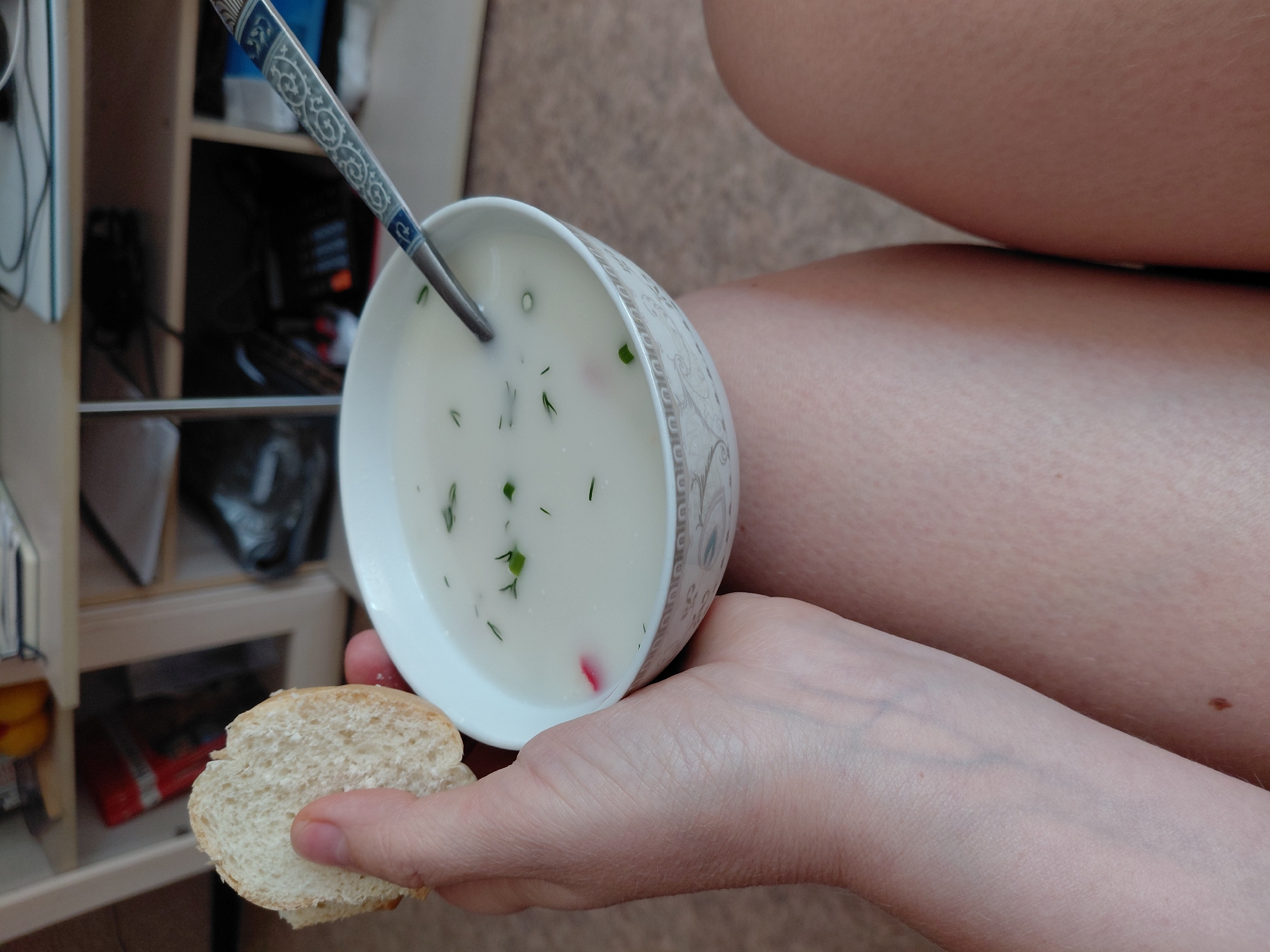 A first-person perspective shot shows a person holding a slice of bread and a bowl of soup. The soup is white with green herbs and small pieces of red and pink ingredients, and a spoon with an ornate pattern is in the bowl. The person's arm and hand are visible in the foreground, and their legs are visible below the soup bowl. The background shows shelves filled with various items, including books, electronics, and what appear to be food items on the top shelf. The scene is indoors, likely in a home environment, and is not identifiable as being in Novosibirsk, Russia.