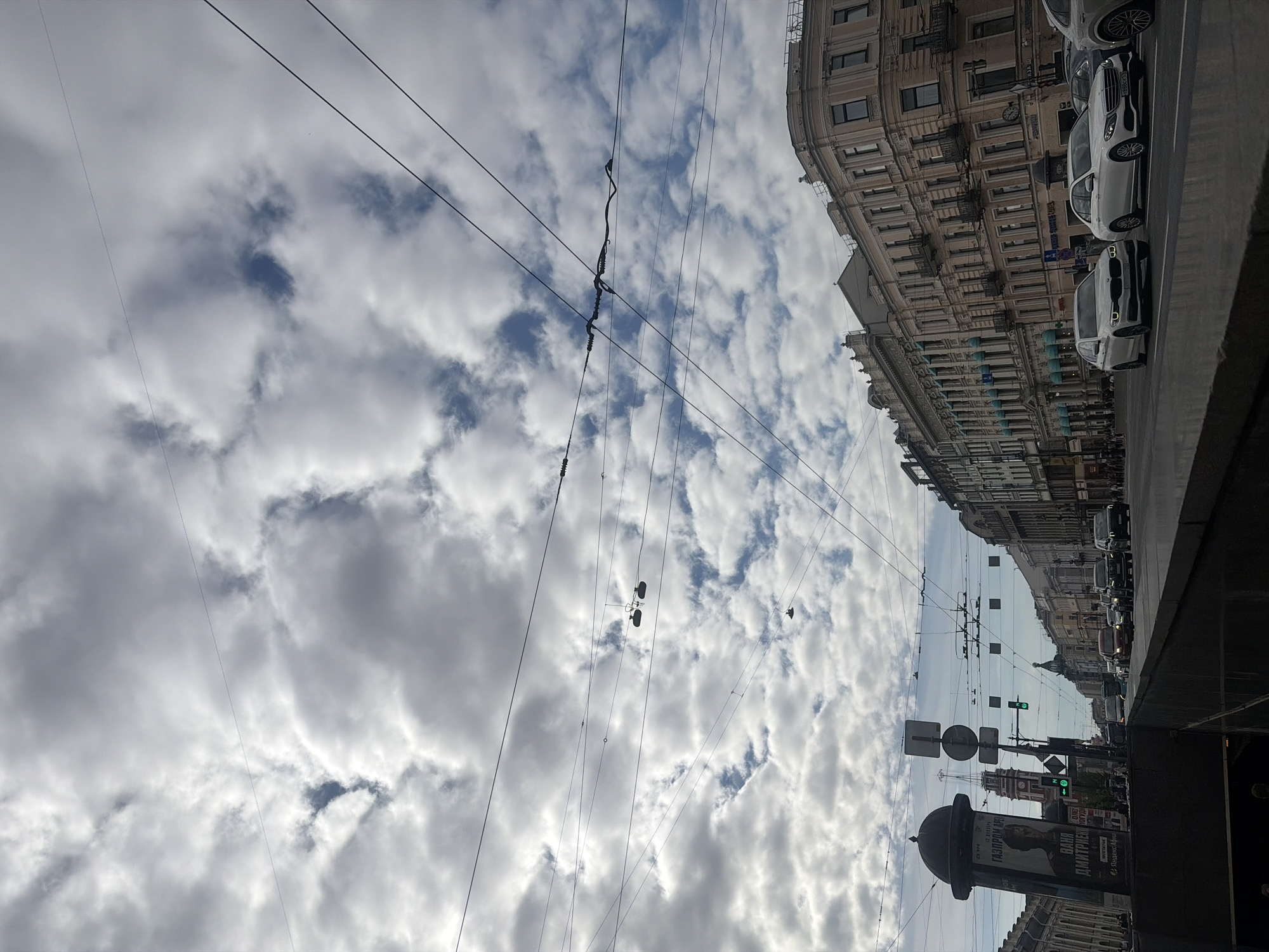 This is a vertical, eye-level, medium shot of a city street in Saint Petersburg, Russia, on a cloudy day. The scene is dominated by a dramatic sky filled with textured, grey and white clouds, broken by patches of blue. Several dark power lines crisscross the sky, creating a graphic, linear element against the organic shapes of the clouds.
The right side of the frame features the ornate architecture of a historic building, with multiple floors of windows and classical detailing. Parked cars, primarily white, line the street in front of the building, suggesting a busy urban environment. Further down the street, traffic is visible, including several dark SUVs and cars, indicating ongoing activity. Streetlights and traffic signals add to the urban landscape.
In the foreground on the right, a dark, textured surface, possibly a building facade or sidewalk, creates a strong vertical line. Towards the bottom center, a large, cylindrical pillar with a domed top stands prominently, adorned with advertisements. One visible advertisement features a portrait of a woman and text that appears to be in Cyrillic. Other street signs and a traffic light showing green are also present.
The time of day is likely late morning or afternoon, given the bright but diffused light filtering through the clouds. The weather is overcast, contributing to the muted color palette and the dramatic sky. The overall impression is one of a bustling city street viewed from a slightly elevated perspective, with the striking sky and classic architecture providing a sense of place. There are no clearly discernible people in the frame, though the presence of cars and advertising suggests a populated area.