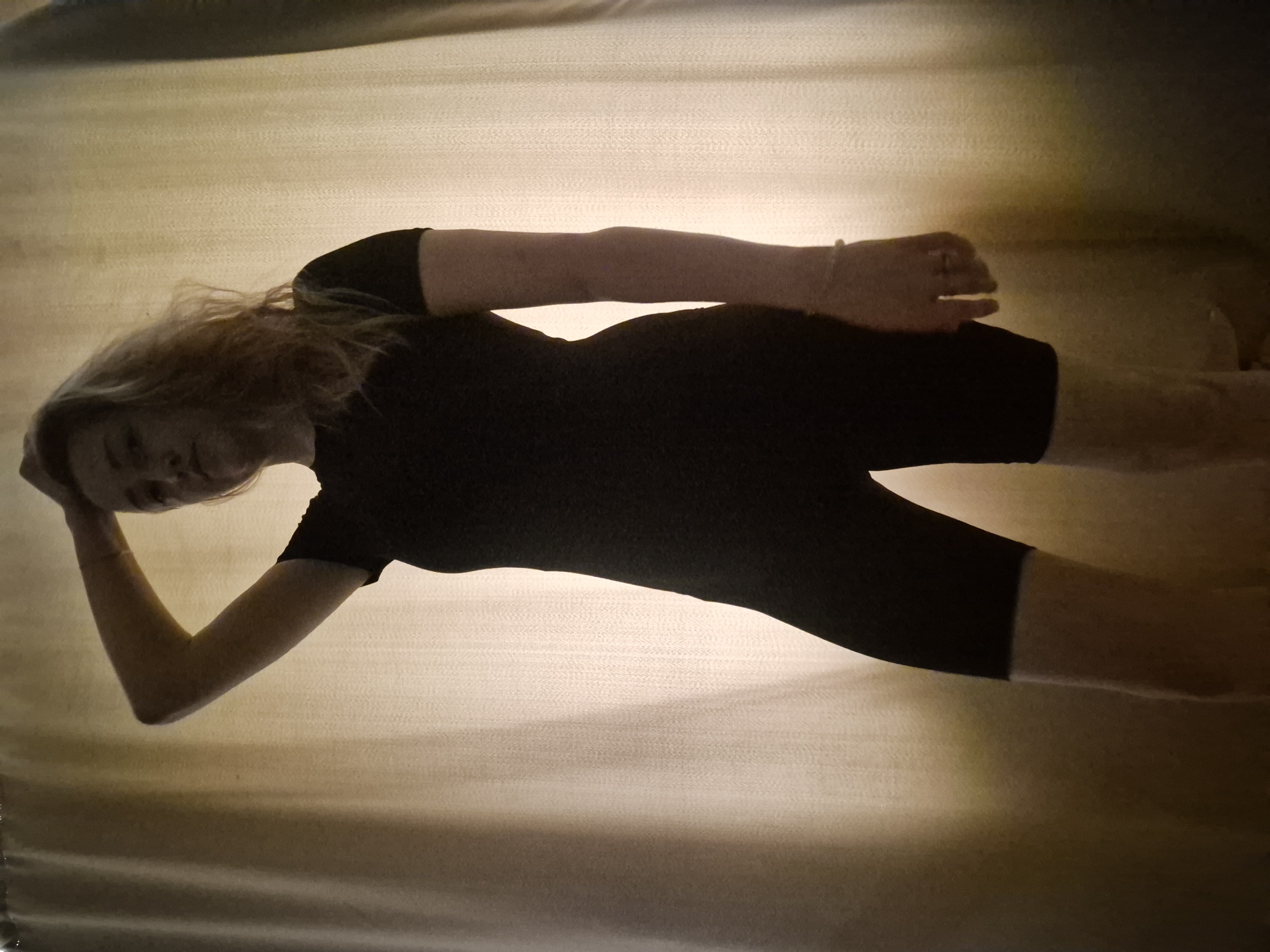 A woman with blonde hair is lying on a beige surface. She is wearing a black bodysuit. Her left arm is bent, and her hand is near her head. Her right arm is extended, and her hand is near her right leg. The location is unknown and there is no evidence to indicate that this image was taken in Chaikovsky, Russia.