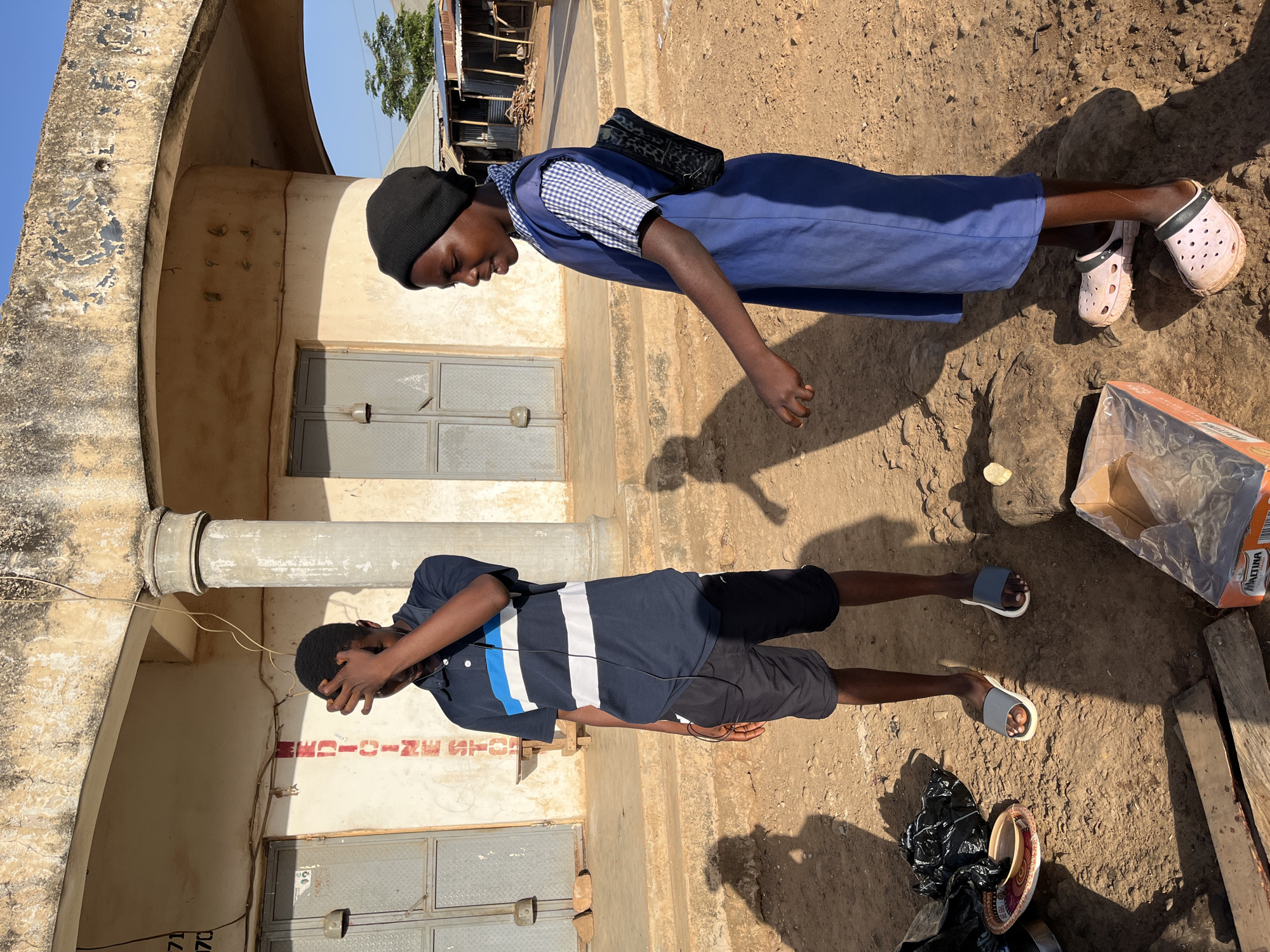 The image depicts two individuals in an outdoor setting, likely in Jos, Nigeria, based on architectural and contextual clues. A young girl wearing a blue school uniform, a black head covering, and pink Crocs stands with her back to a wall of a building. She wears a patterned bag over her shoulder.
In the foreground, a young man in a dark blue polo shirt with white and blue stripes, black shorts, and gray/white sandals is standing next to the same building. He has his right hand on his forehead, seemingly shielding his eyes.
The setting is an outdoor dirt area with scattered debris, including what appears to be packaging materials and trash. The building facade is light-colored and shows visible wear and tear. The presence of doors suggests an entry way or shop front. The scene appears to be in daylight.