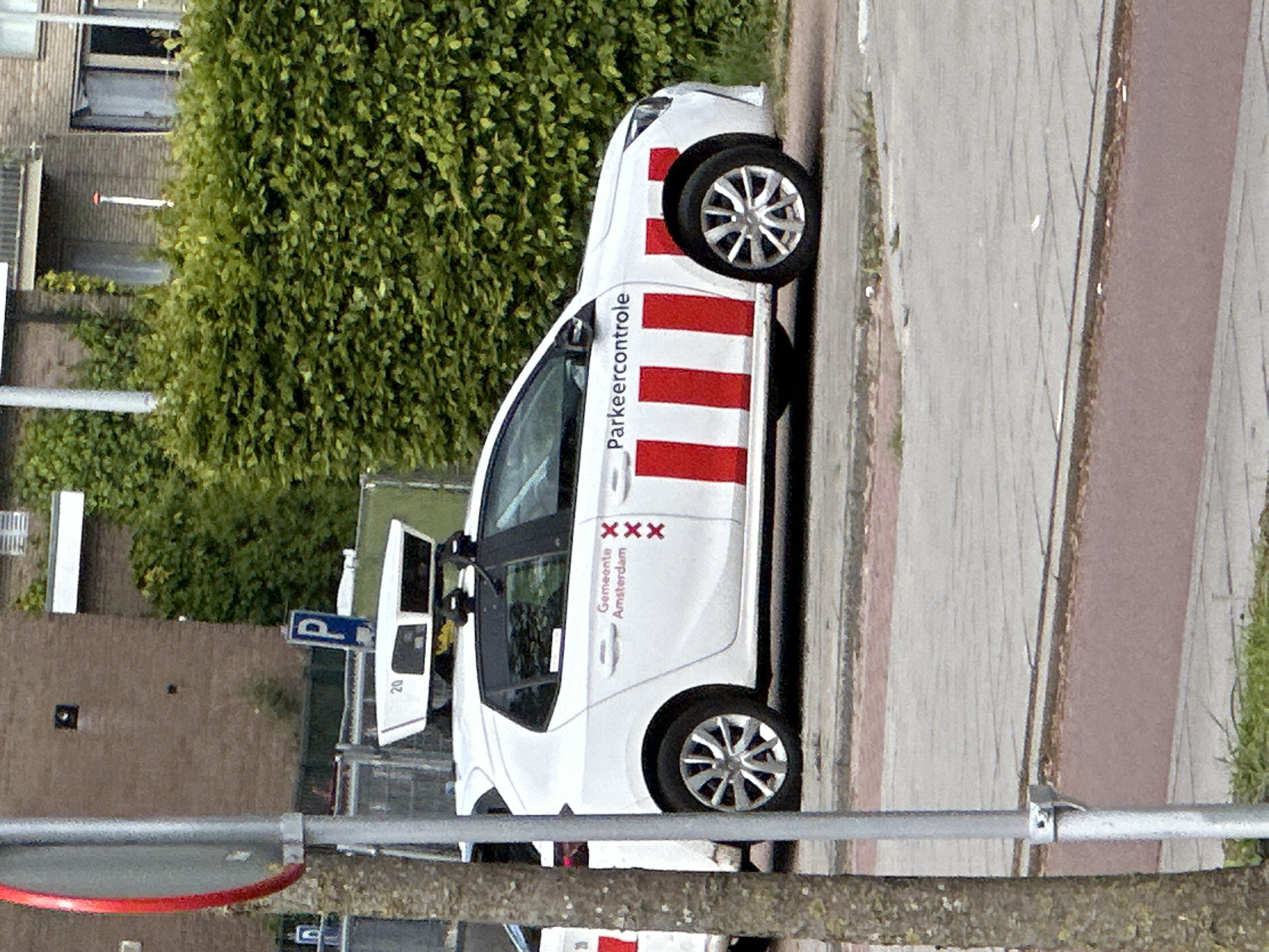 The image shows a white car parked on the side of a street in Amsterdam, Netherlands. The car has red and white stripes along its side and "Parkeercontrole" (Parking Control) and "Gemeente Amsterdam" (Municipality of Amsterdam) written on it. The Amsterdam coat of arms is visible. The car appears to be a parking enforcement vehicle. A parking sign is visible nearby. The surroundings include a sidewalk, a building, and greenery.
