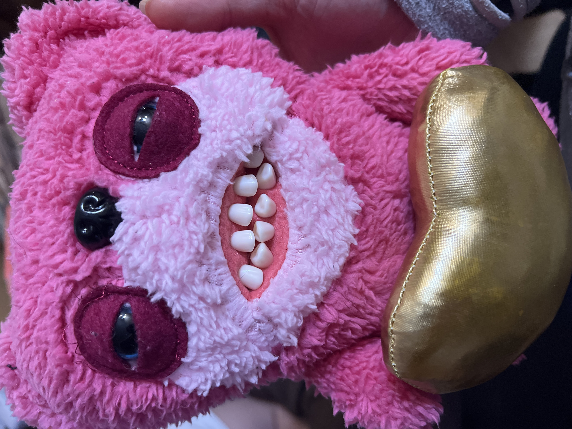 A pink plush toy is held up in someone's hand. The toy has three eyes and a mouth containing a set of small, white teeth. A gold-colored bean-shaped object is attached to the side of the toy. The location is not discernible from the image. No people are visible beyond the hand holding the toy.