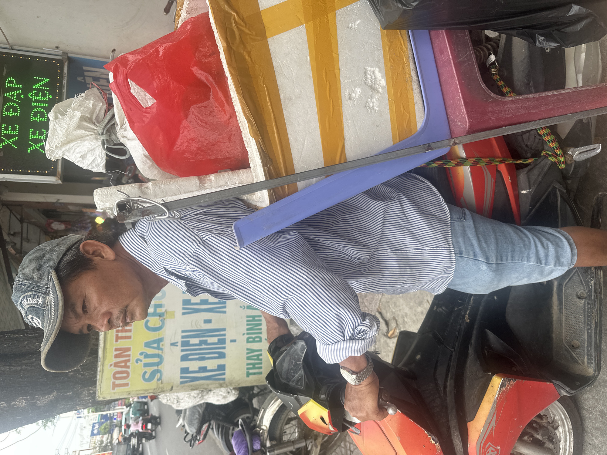 The scene, occurring in Bac Nha Trang Ward, Vietnam, features a man on a red motorcycle. He is wearing a gray baseball cap and a blue striped shirt, with the sleeves rolled up. A watch is visible on his wrist as he grips the motorcycle's handlebars.
The motorcycle is laden with various items, including a red plastic bag, a white styrofoam block secured with yellow tape, and a purple plastic chair. A black plastic bag is also present among the items.
Behind the man and motorcycle is a sign with Vietnamese writing, part of which is in green LED lights. The sign appears to advertise repair services for electric vehicles. Further in the background, a streetscape with other motorcycles and vehicles is visible.