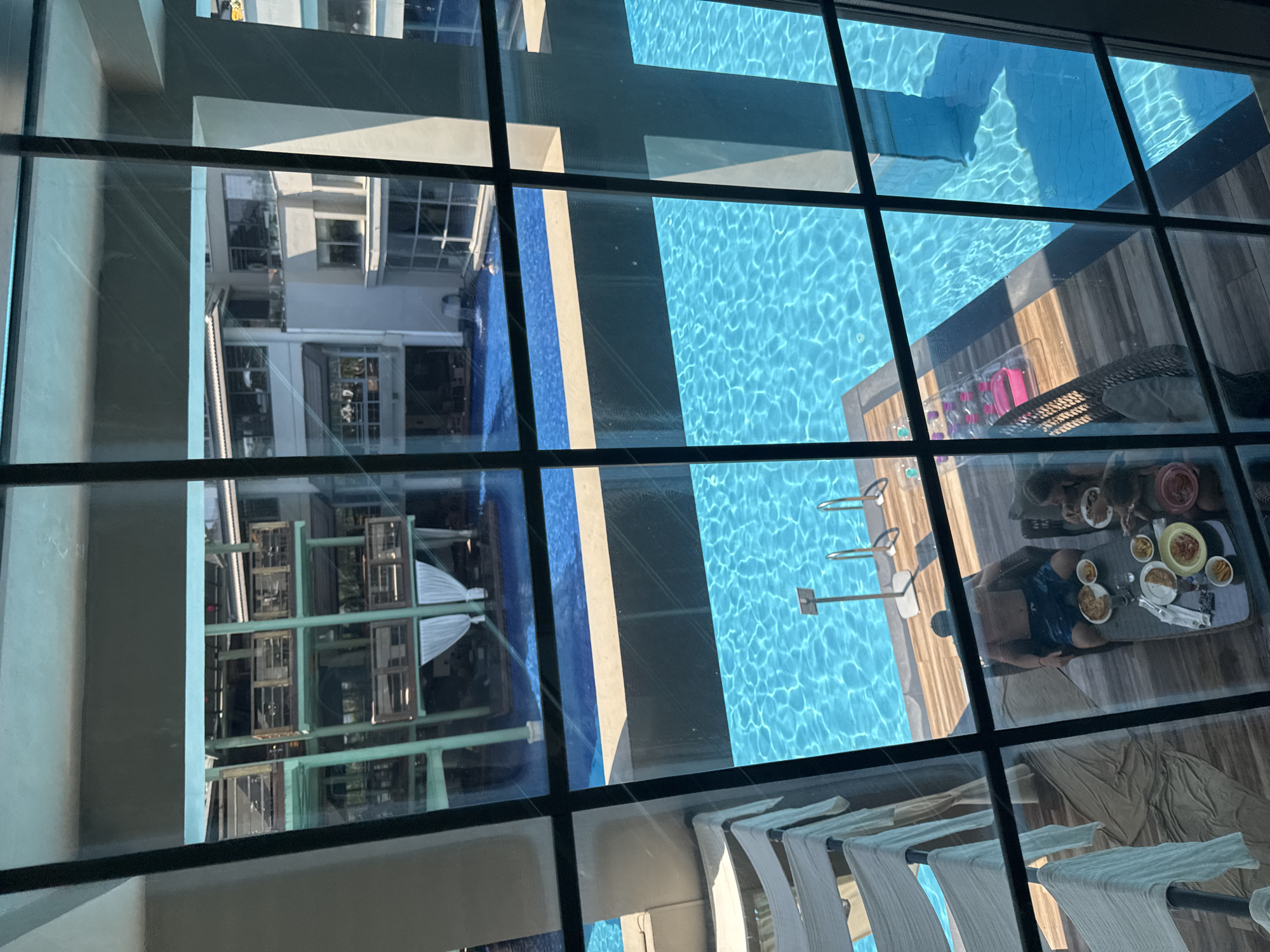 The image shows a view of a swimming pool area, likely in a resort or hotel, with buildings in the background. The pool is a prominent feature, with bright blue water reflecting sunlight. In the foreground, a group of people are seated around a table on a wooden deck, appearing to be eating. There are lounge chairs and white fabric structures that suggest a shaded seating area. The overall scene depicts a relaxing, sunny environment. The image is taken from an elevated position, looking down into the pool area, and is partially obscured by window panes. The context provided suggests this scene is located in Aksu, Turkey.