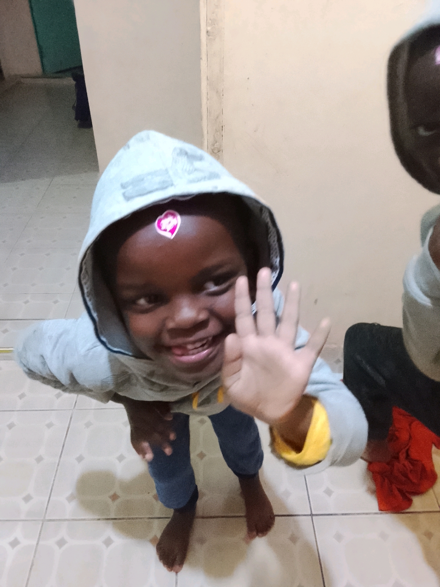 A medium shot shows a young child with dark skin and wearing a gray hooded sweatshirt with a pink heart sticker on their forehead. The child is smiling and holding up their right hand, showing four fingers. The child is wearing blue jeans and standing on a tiled floor. The background is a cream-colored wall. Another child, out of focus and wearing a dark shirt, is visible to the right. A red piece of fabric is on the floor next to the second child. The scene appears to be indoors, possibly in a residential setting in an unknown location.
