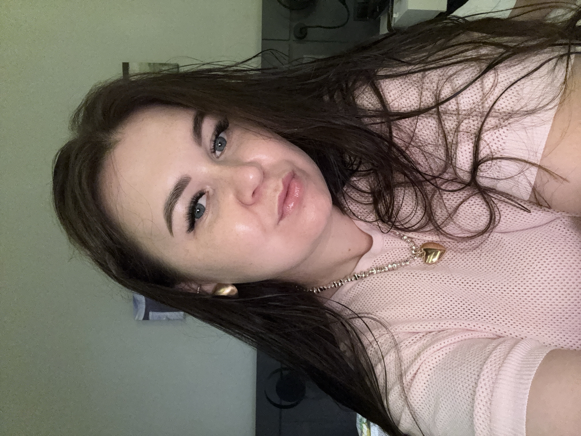 The image shows a close-up, head-and-shoulders selfie of a woman with long, dark brown hair, blue eyes, and fair skin with freckles. She is wearing a light pink, textured, short-sleeved sweater and a gold necklace with a heart-shaped pendant. She has on dramatic false eyelashes and her lips appear glossy. The background is dimly lit and appears to be an indoor setting, possibly a bedroom, with a gray wall and what looks like a headboard of a bed visible behind her. There is also some electronic equipment and possibly a picture on the wall in the background. The lighting suggests an artificial light source, possibly from above and to the front. There are no recognizable landmarks or objects that would indicate the location is Alanya, Turkey.
