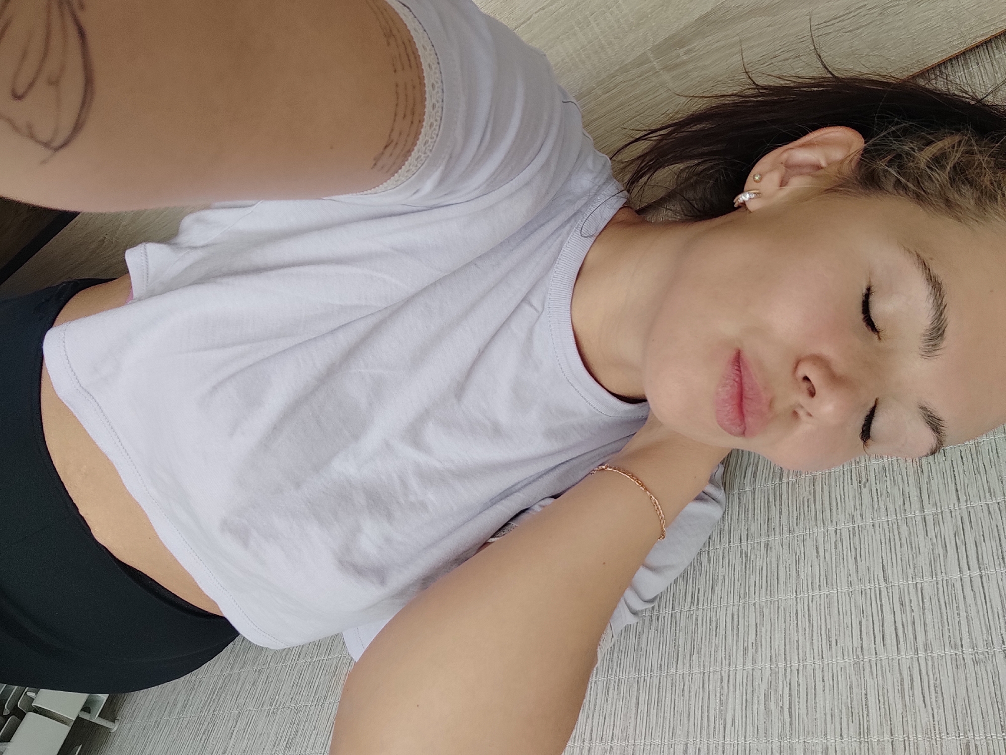 A young woman is captured in a high-angle shot, lying relaxed on a textured light-colored mat indoors in Chaikovsky, Russia. Her body is positioned diagonally across the frame, with her head resting on the upper right side. Her eyes are closed, indicating a state of rest, relaxation, or sleep, and her expression appears serene. Her dark hair is spread loosely around her head.
She is dressed in a light-colored, possibly pale grey or white, cropped t-shirt that reveals a sliver of her midriff, and black bottoms, likely leggings or shorts. A delicate gold-colored chain bracelet is visible on her right wrist. Her left ear features two small earrings: a stud in the lobe and a small hoop with clear stones in the cartilage.
Notable details include markings on both arms. On her left upper arm/shoulder, there is a distinct outline of a drawing or tattoo, resembling a butterfly or an abstract winged shape. On her right upper arm, several thin, concentric ring-like lines or a tattoo are visible. The background consists primarily of the textured mat, which features a repeating pattern of parallel lines, and a segment of wooden flooring on the left and top edges of the image. The overall lighting is soft and even, suggesting a calm indoor setting.