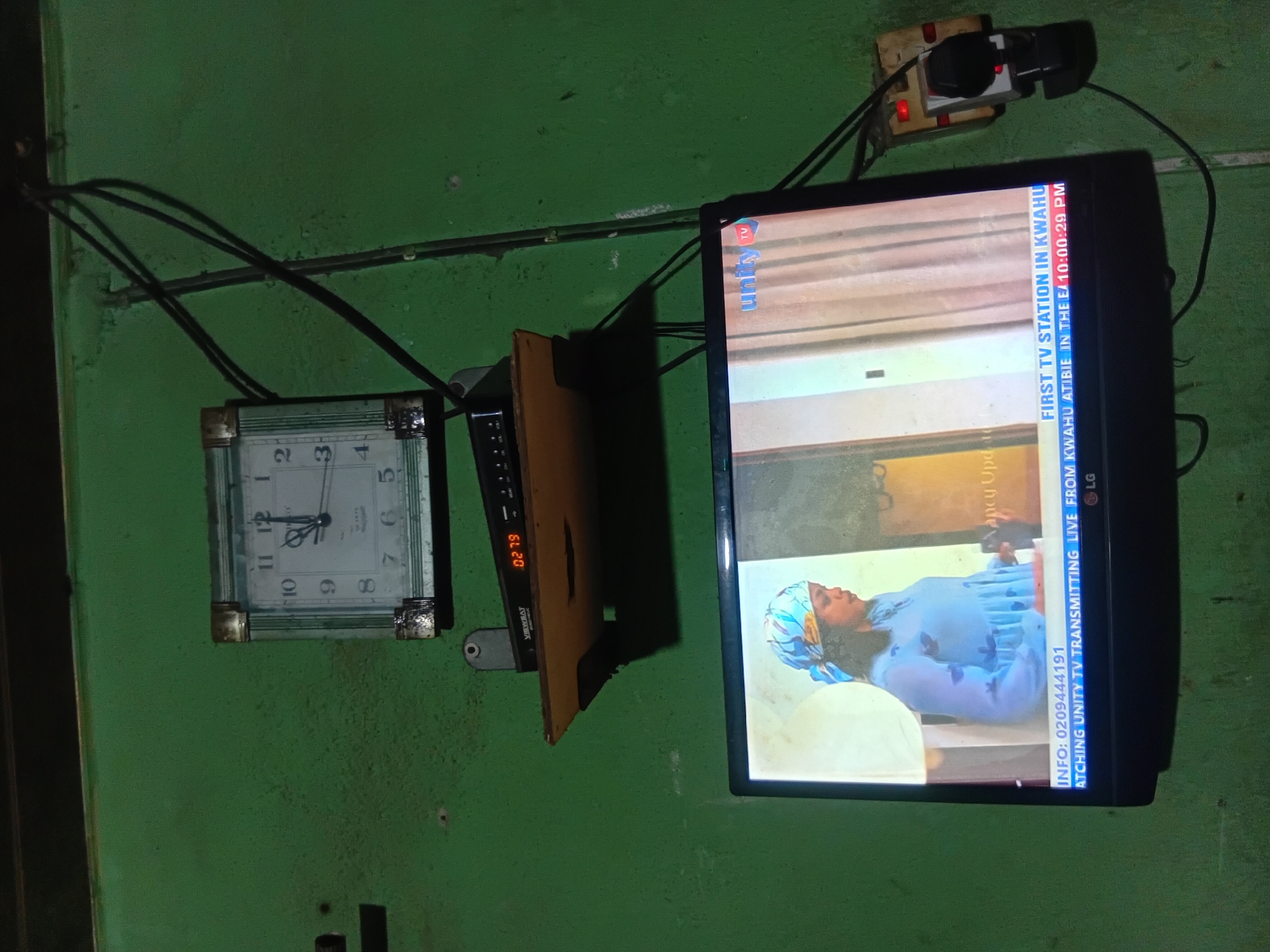 The image shows a wall in Nkpor, Nigeria, with a square clock on the upper part, a shelf with a set-top box below it, and a television at the bottom. The clock is a white analog clock with Roman numerals and a metallic border. The set-top box is black with an orange digital display showing "02.79". The television is displaying a news program or a show with a woman in a blue dress and headscarf. The lower part of the television screen has text overlays. The wall is painted green and appears textured with some signs of wear. There are black electrical wires running along the wall and connected to a power socket with a plug. The overall scene depicts a typical domestic setting in a Nigerian home.