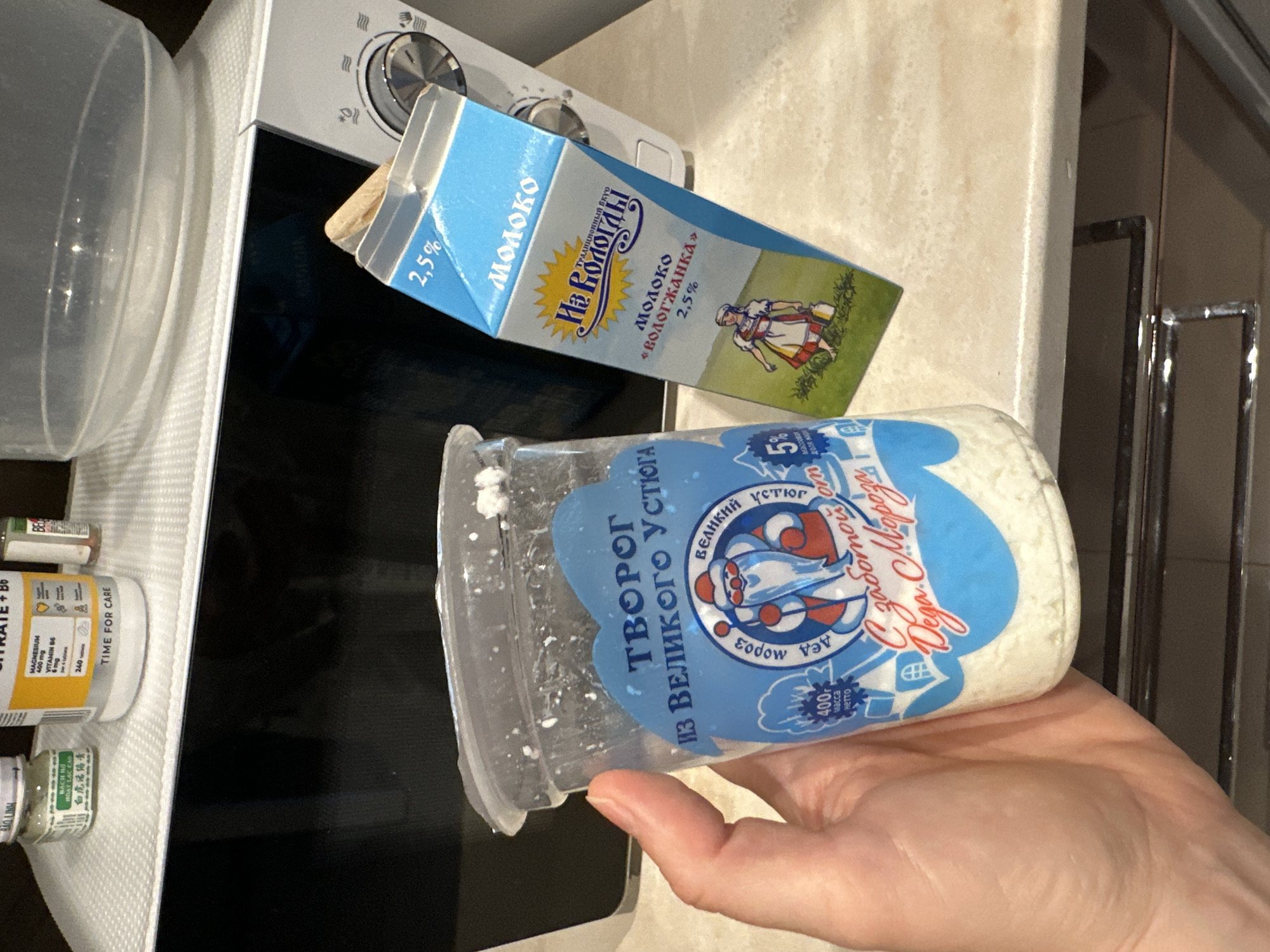 A person is holding a clear plastic container of cottage cheese in front of a microwave oven. The cottage cheese is white and crumbly and fills the bottom half of the container. The container has a blue label with text in Russian that reads "ТВОРОГ ИЗ ВЕЛИКОГО УСТЮГА" (Tvorog iz Velikogo Ustyuga), which translates to "Cottage Cheese from Veliky Ustyug." There is also an illustration of Grandfather Frost on the label. To the right of the cottage cheese container is a blue and white carton of milk with the brand name "МОЛОКО" (Moloko) in large letters. The milk carton has a picture of a woman in traditional clothing. The microwave is white and has a black door. There are also some vitamin bottles visible in the background on top of the microwave. The scene is indoors, on a kitchen counter. The lighting suggests it is daytime.