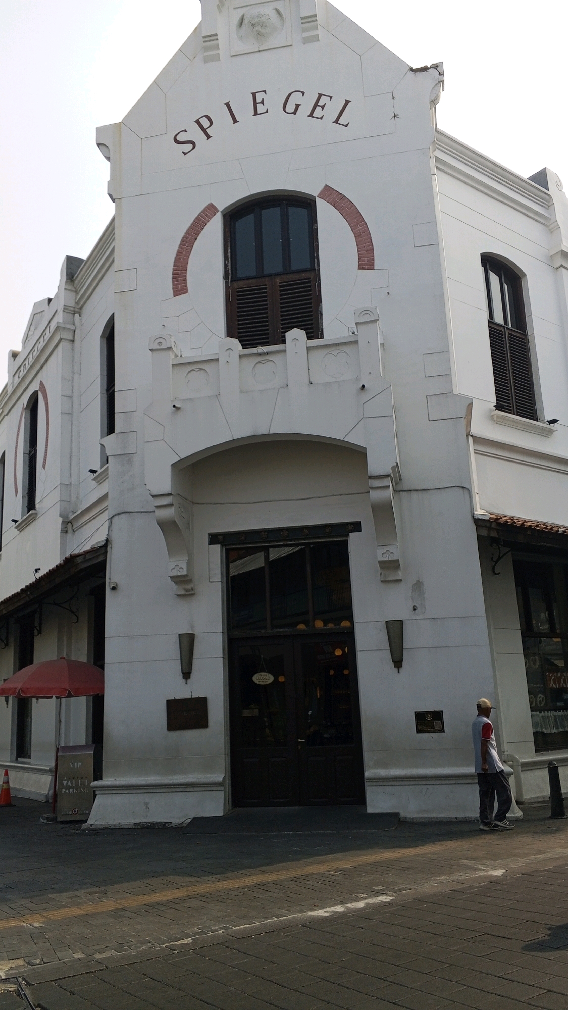 The image captures the Spiegel Building, a historic landmark located in Semarang, Indonesia, a city on the island of Java. The building exhibits colonial architecture, with a white facade, arched windows, and decorative elements. Above the main entrance, the name "SPIEGEL" is inscribed in large, dark red letters. The building has multiple stories, with dark-shuttered windows on the upper levels.
In front of the building, a red umbrella stands to the left, adjacent to a sign that reads "VIP VALET PARKING." An orange traffic cone is also visible near the umbrella. The ground in front of the building is paved with dark cobblestones.
A man wearing a white cap, a red and white shirt, and grey pants is walking away from the building on the right side of the image. He appears to be strolling along the sidewalk. The scene suggests a daytime setting, with the sun casting shadows on the pavement. The overall atmosphere is one of historical preservation and urban life.