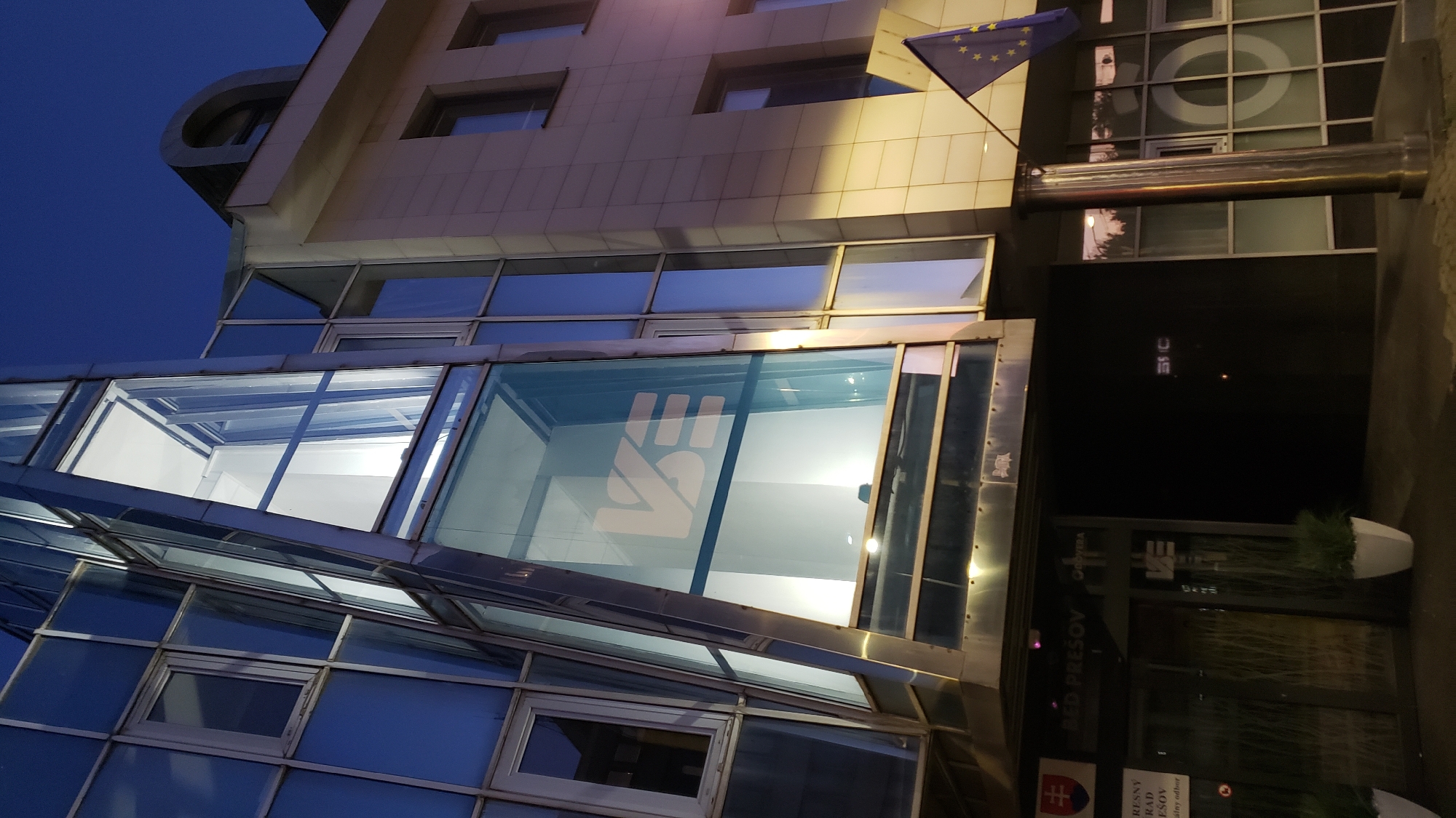 The image shows a modern building at dusk in Prešov, Slovakia. The building features a prominent glass facade, with a section angled upwards, revealing internal lighting. A large logo, possibly "VSE", is visible on this glass section. To the right, a section of the building is clad in light-colored stone, with several windows. A European Union flag flies from a pole in front of this section. Below the glass facade, at ground level, there is an entrance with signage indicating "BED PREŠOV". Planters with greenery are positioned near the entrance. The scene is set against a dark blue sky, suggesting it is either early evening or late twilight.