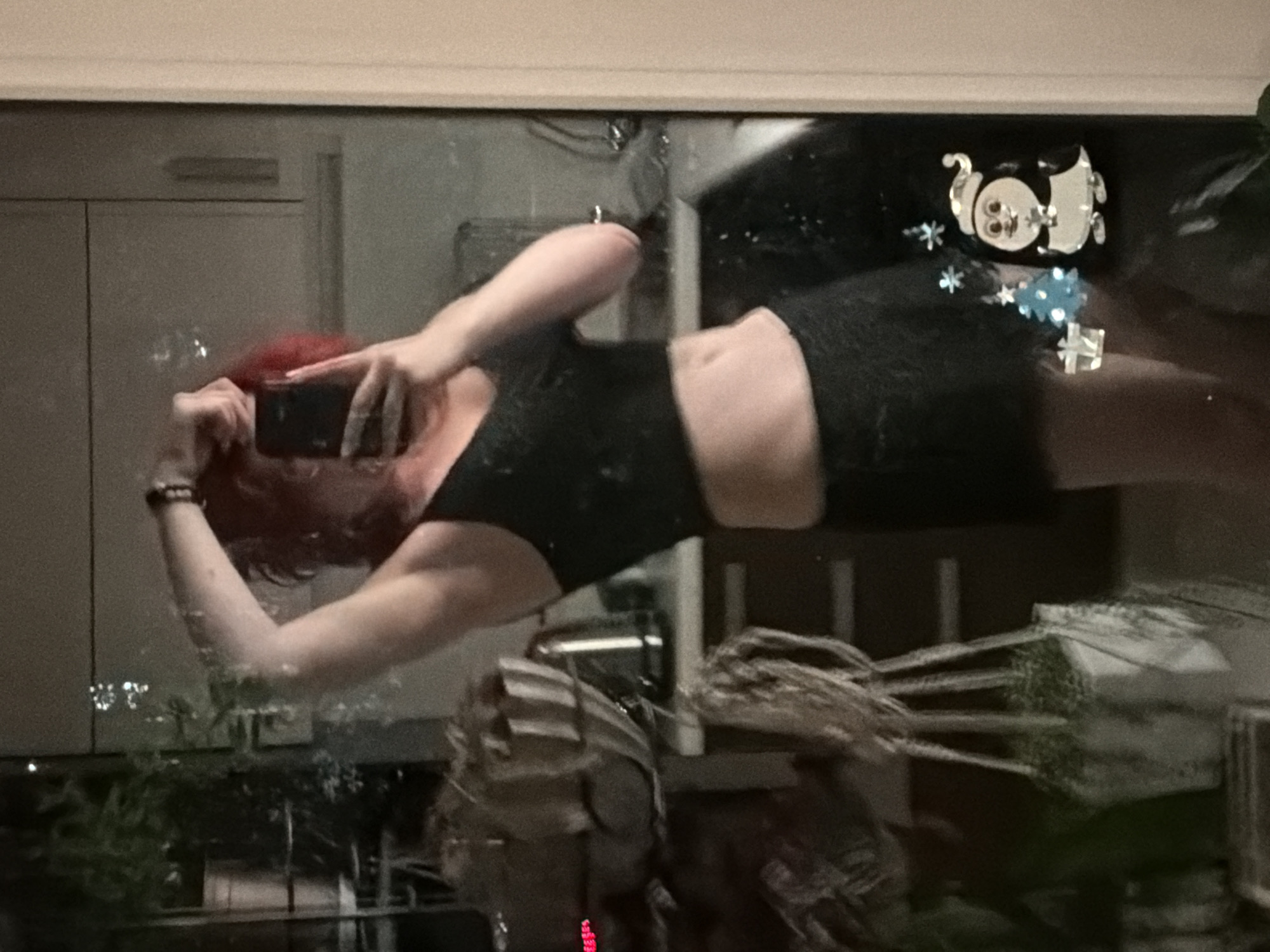A person with red hair is taking a mirror selfie in what appears to be a bathroom or kitchen. The person is wearing a black crop top and black shorts. Their right arm is raised, and they are holding a smartphone to take the picture. In the background, there are reflections of plants, kitchen cabinets, and a kettle. There are also Christmas-themed stickers on the mirror, including a penguin, a Christmas tree, and a gift. The scene is dimly lit, and the image quality is somewhat blurry. The location is not specified within the image or its description.