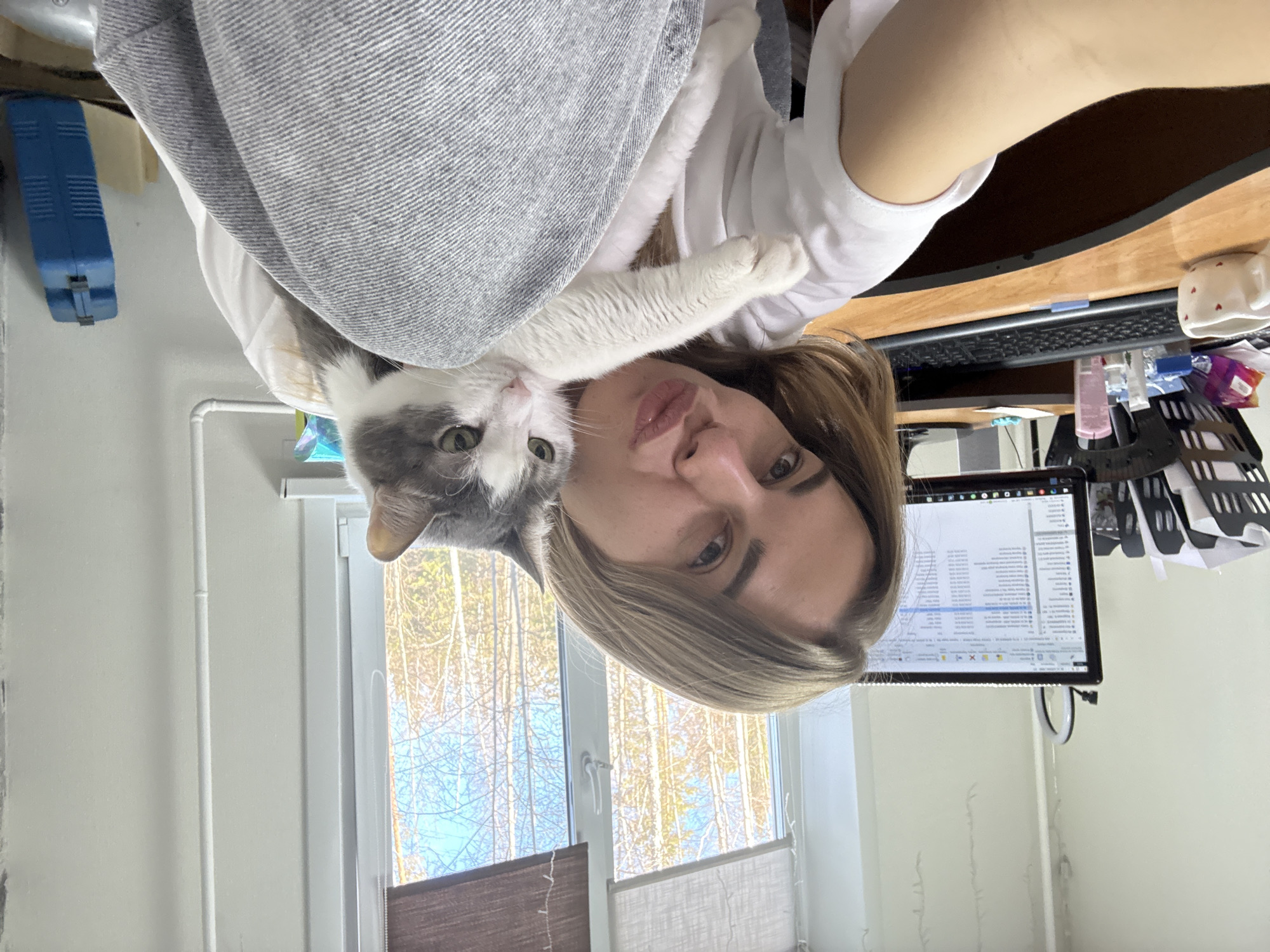 The image, originally rotated 90 degrees clockwise, presents a close-up selfie of a person and a cat in an indoor setting.
**Subjects:** The primary subjects are a person, likely female, with light brown hair, and a domestic cat. The person looks directly at the camera with a neutral expression, featuring full lips and light-colored eyes. They are wearing a white t-shirt, partly covered by a light grey textured fabric, possibly a blanket. The cat, with distinctive grey and white fur and striking green eyes, is resting on the person's chest or shoulder, also looking towards the camera, with its front paws draped over the person's arm.
**Scene and Setting:** The setting appears to be a home office or a room with a workspace. To the person's left (when correctly oriented) is a large window revealing an outdoor scene. The trees beyond the window are bare, suggesting a cold season such as late autumn, winter, or early spring in Ust-Ilimsk, Russia, under a bright blue sky. On the person's right, a wooden desk is visible, holding a computer monitor displaying what appears to be a spreadsheet or a document filled with text in columns, indicative of data or a list. A keyboard sits in front of the monitor. Other desk items include a white mug adorned with small red heart patterns, a black wire basket filled with papers, and several small bottles or containers. A blue, rectangular wall-mounted item is seen behind the desk.
**Activity:** The person is taking a selfie while holding or cuddling the cat.
**Notable Details:** The bright light from the window indicates it is daytime. The scene conveys a calm and personal moment. The visible outdoor environment, with bare trees, is consistent with the location context of Ust-Ilimsk, Russia.
**Visible Text:** The computer screen displays rows and columns of text, suggesting a software application such as a spreadsheet program. Standard window control buttons (minimize, maximize, close) are visible on the application title bar on the monitor. No specific words or phrases are clearly legible due to the distance and resolution.