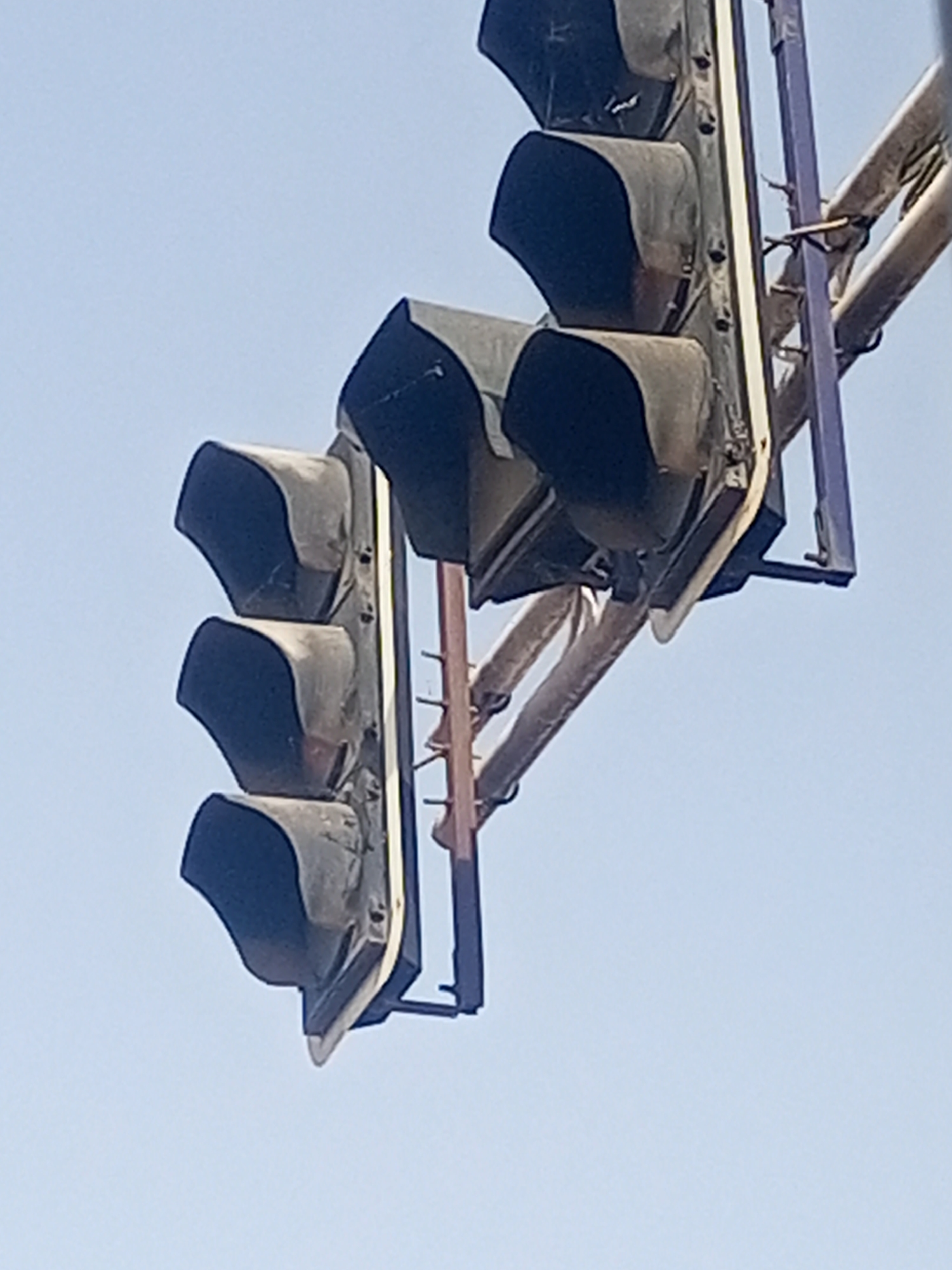 The image captures two sets of traffic lights against a clear, light blue daytime sky in Maiburiji, Nigeria. On the left, a set of three vertically stacked traffic light units is visible. Each unit is dark-colored, likely black or dark gray, and equipped with a deep visor or hood designed to shade the light from direct sunlight. A second, partially seen set of traffic lights appears on the right, also showing dark-colored units with visors, suspended from an overhead structure.
Both traffic light assemblies are attached to support frameworks. These include light-colored vertical and horizontal elements, possibly metal poles or frames, and a more prominent vertical bar with a distinct purple or dark blue hue connecting to the rightmost traffic light unit. The visible components of the traffic lights and their supports appear somewhat weathered or dusty, consistent with outdoor infrastructure. The sky is bright and free of clouds, indicating sunny weather. No people, vehicles, or specific activities are depicted. There is no visible text on any of the objects in the image. The traffic lights do not appear to be illuminated.