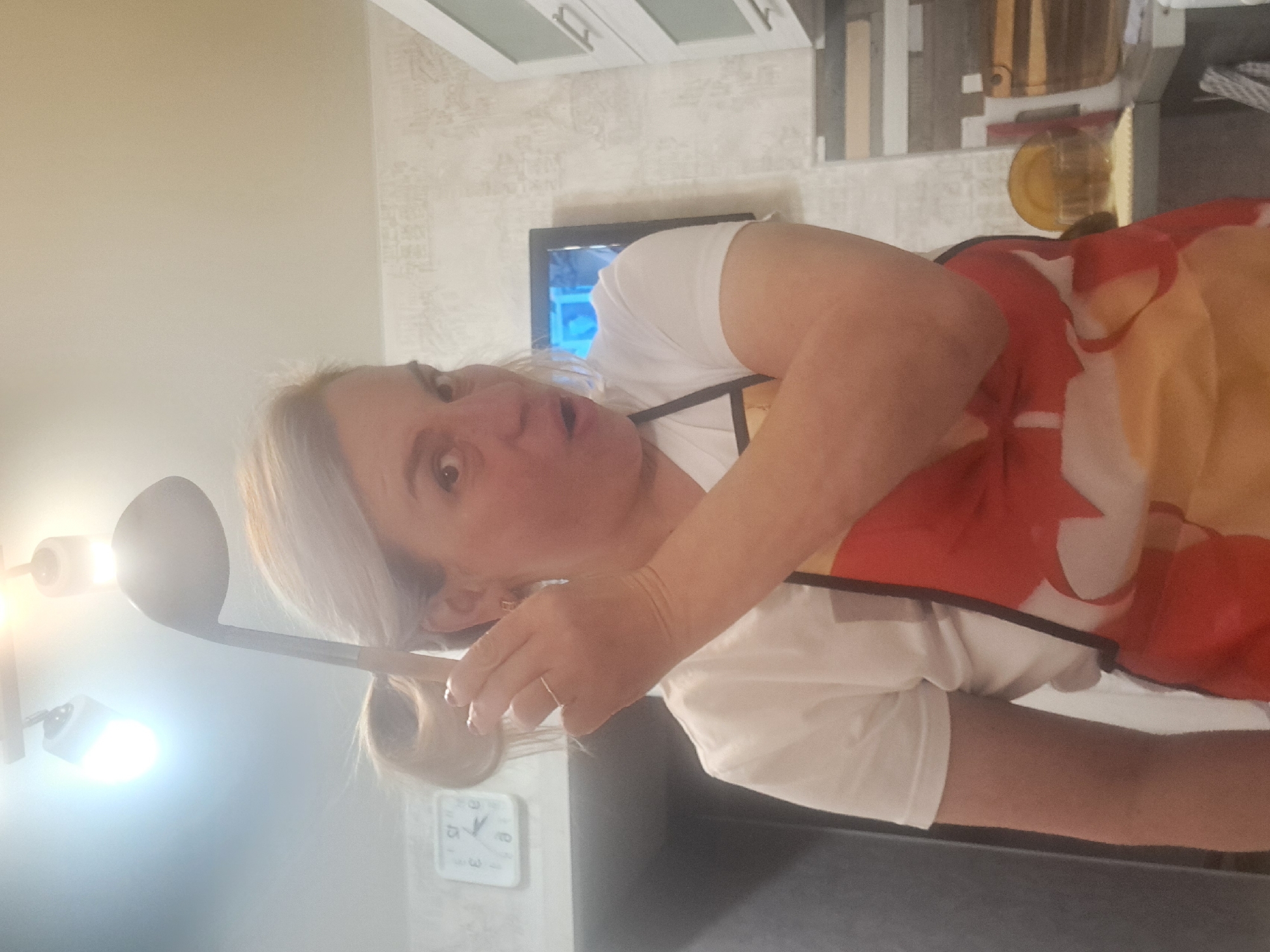 A middle-aged woman with blonde hair, wearing a white t-shirt and a red and yellow apron, is holding a ladle. She appears to be in a kitchen, evidenced by the presence of cabinets, a television, and a clock on the wall. The specific location within the city of Elektrostal, Russia, cannot be determined from the given image.