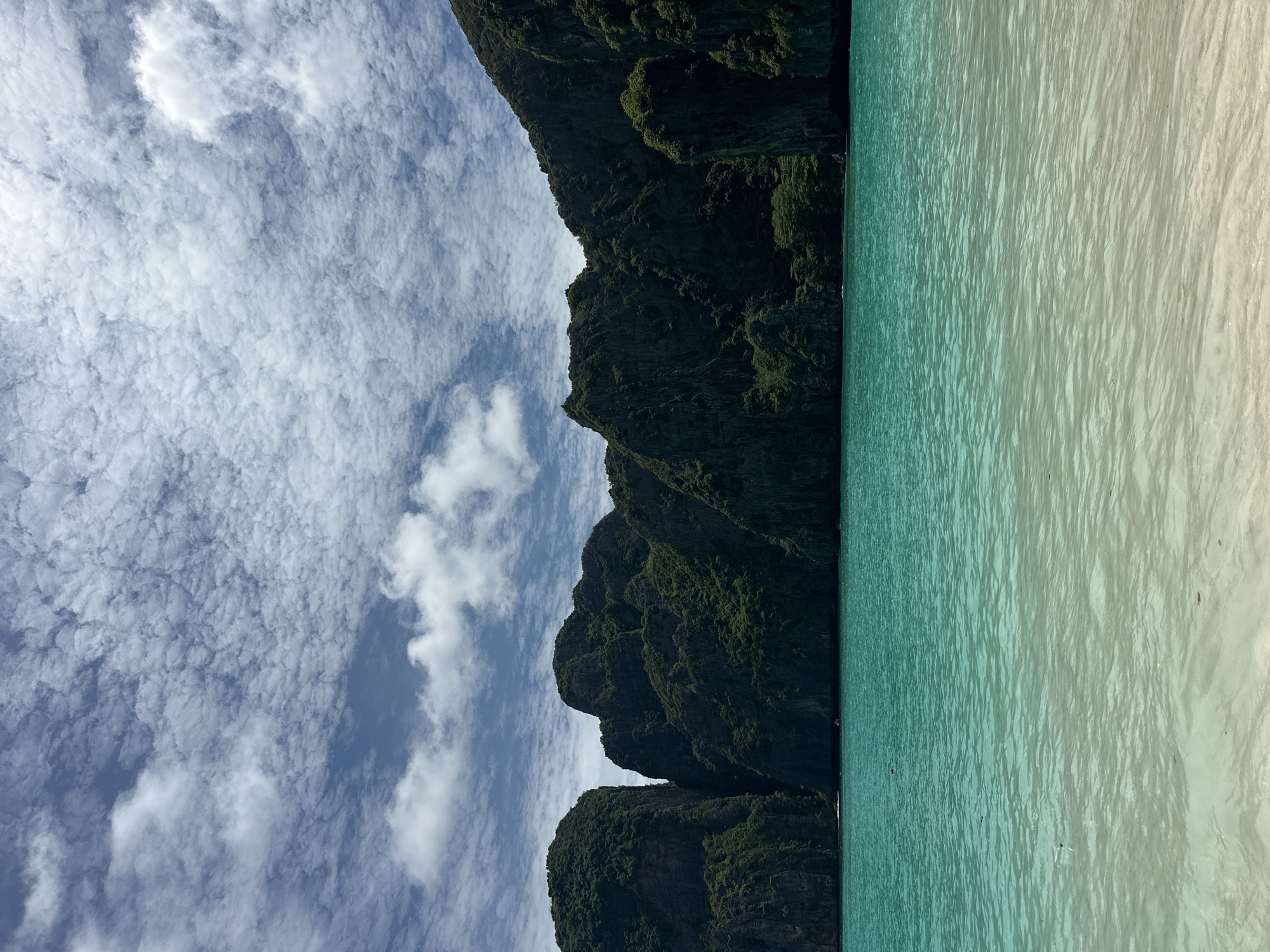 The image depicts a serene tropical beach scene, likely in Ao Nang, Thailand. In the foreground, a shallow, calm turquoise sea gently laps onto a sandy beach. The water transitions from a pale, almost translucent hue near the shore to a deeper, vibrant turquoise further out. In the mid-ground, a line of majestic, jungle-covered limestone karsts rises dramatically from the sea, their dark, rugged faces contrasting with the bright water and sky. The karsts are densely packed with lush green vegetation, hinting at a rich ecosystem. The sky above is a brilliant blue, adorned with a captivating pattern of wispy, white cirrocumulus clouds that create a textured, dappled effect. The overall impression is one of natural beauty and tranquility. There are no visible people or specific man-made structures in this particular frame, emphasizing the untouched, natural landscape.