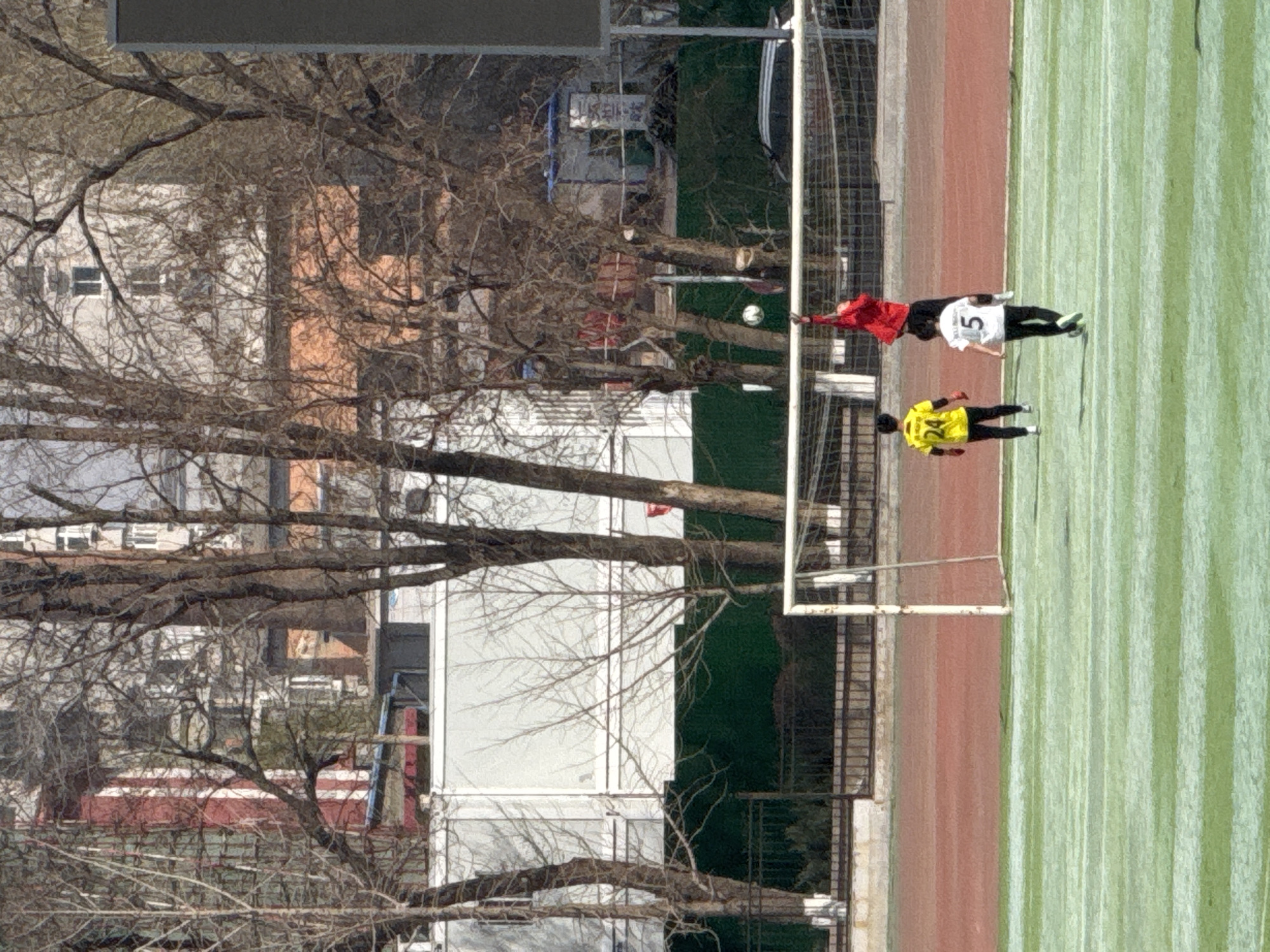 A soccer game is in progress on a striped green field near a red running track in Zhongtun, China. Two players wearing soccer uniforms stand near the goal, one in yellow and black with the number 24 and the other in white and black with the number 5. A red-jacketed person stands close by. A soccer ball floats in the air between them. Trees are in the foreground with several buildings in the background.